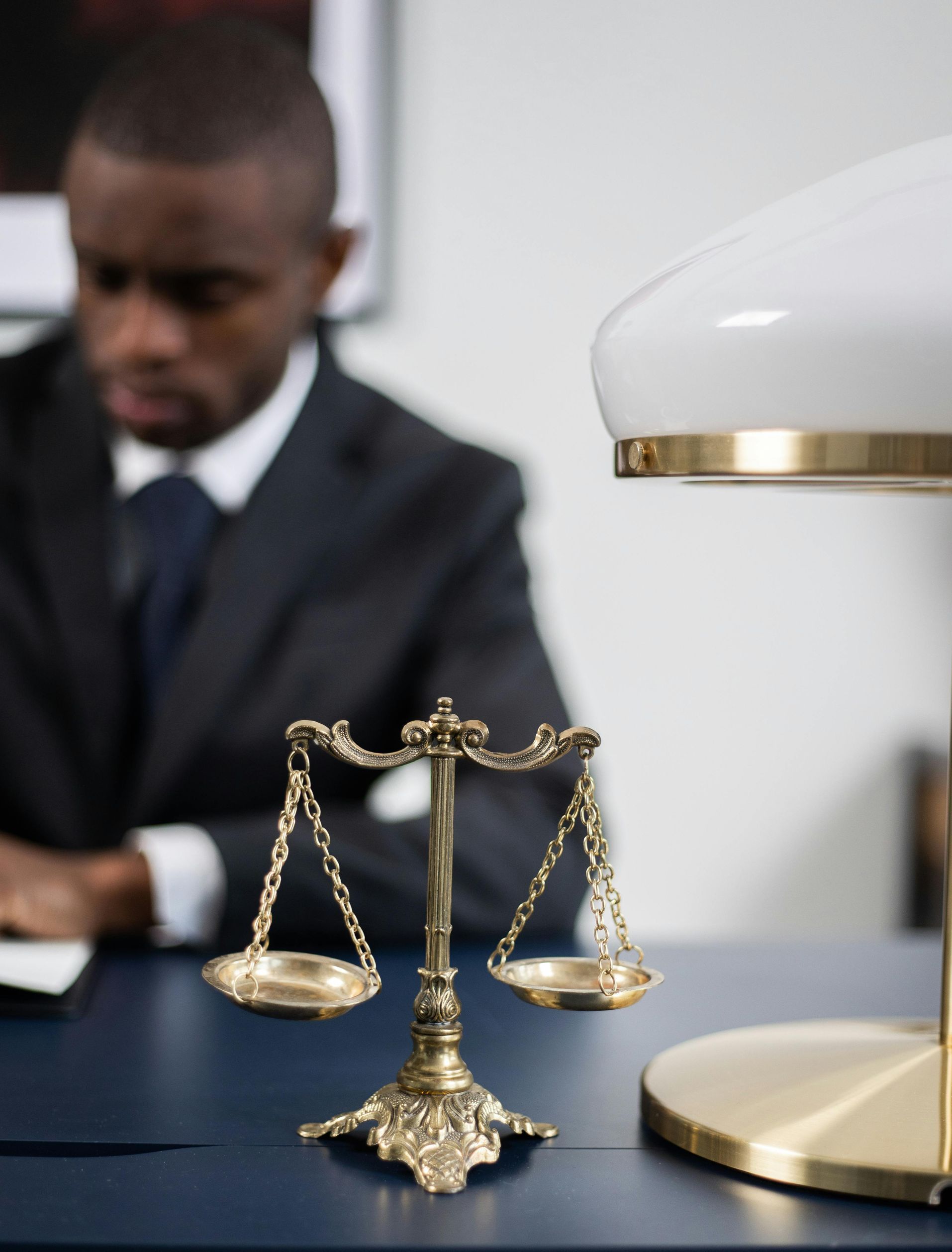 A scale of justice sits on a desk in the foreground, with a professional in a suit working in the blurred background.