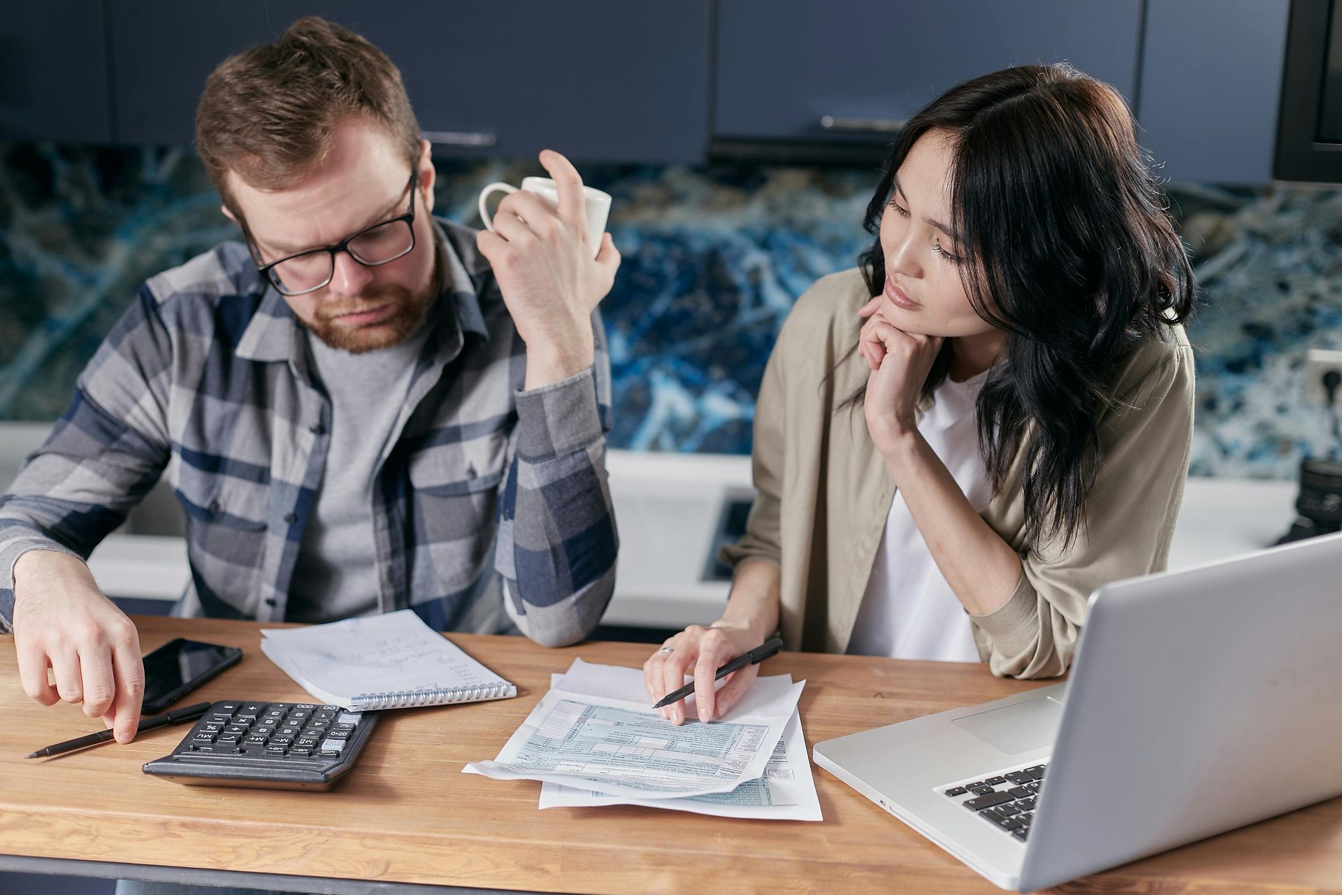 Two people sit at a wooden table reviewing financial documents with a calculator and laptop in a modern kitchen.