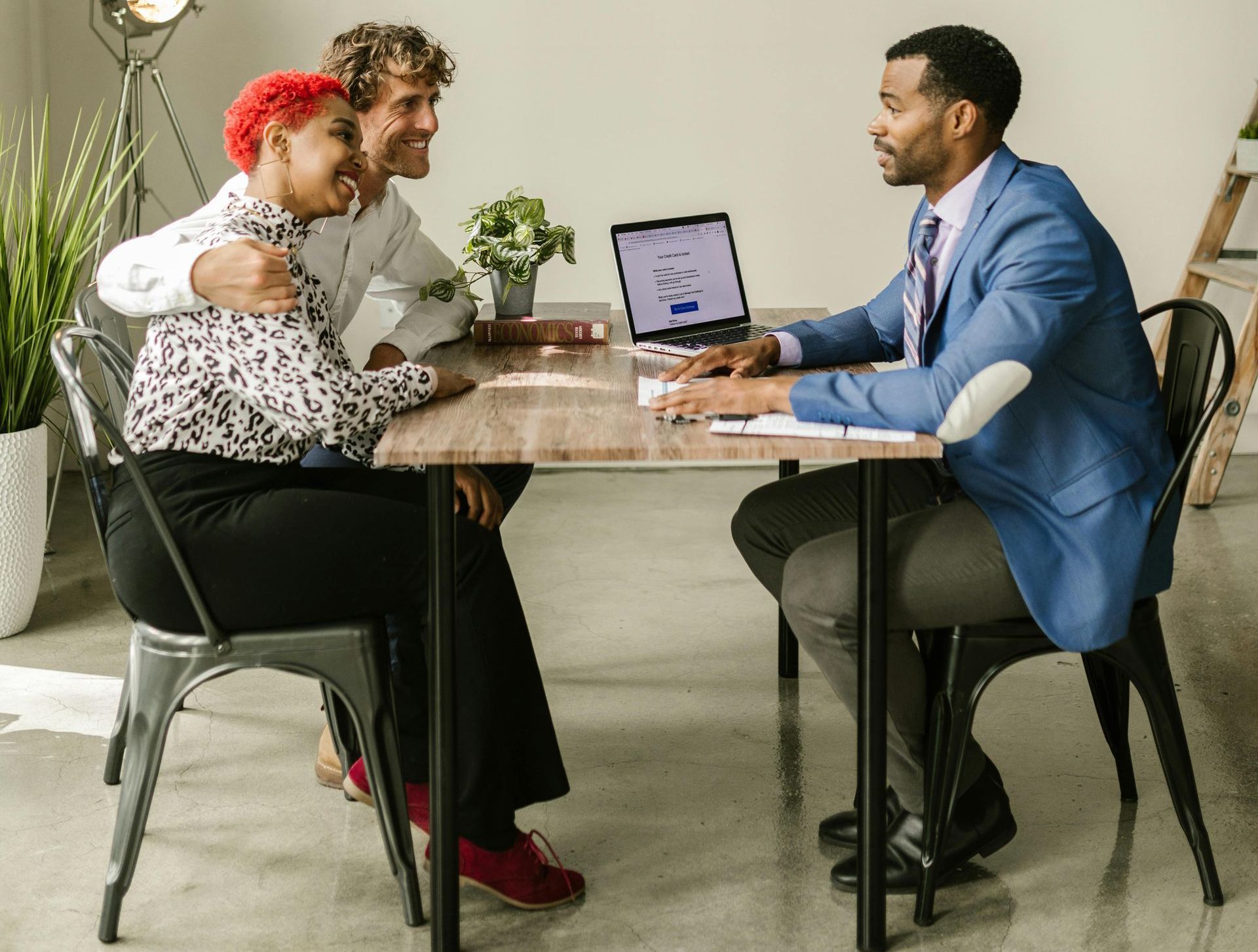 Two people sit at a desk smiling at a professional in a blue blazer during a meeting in a bright, modern office.
