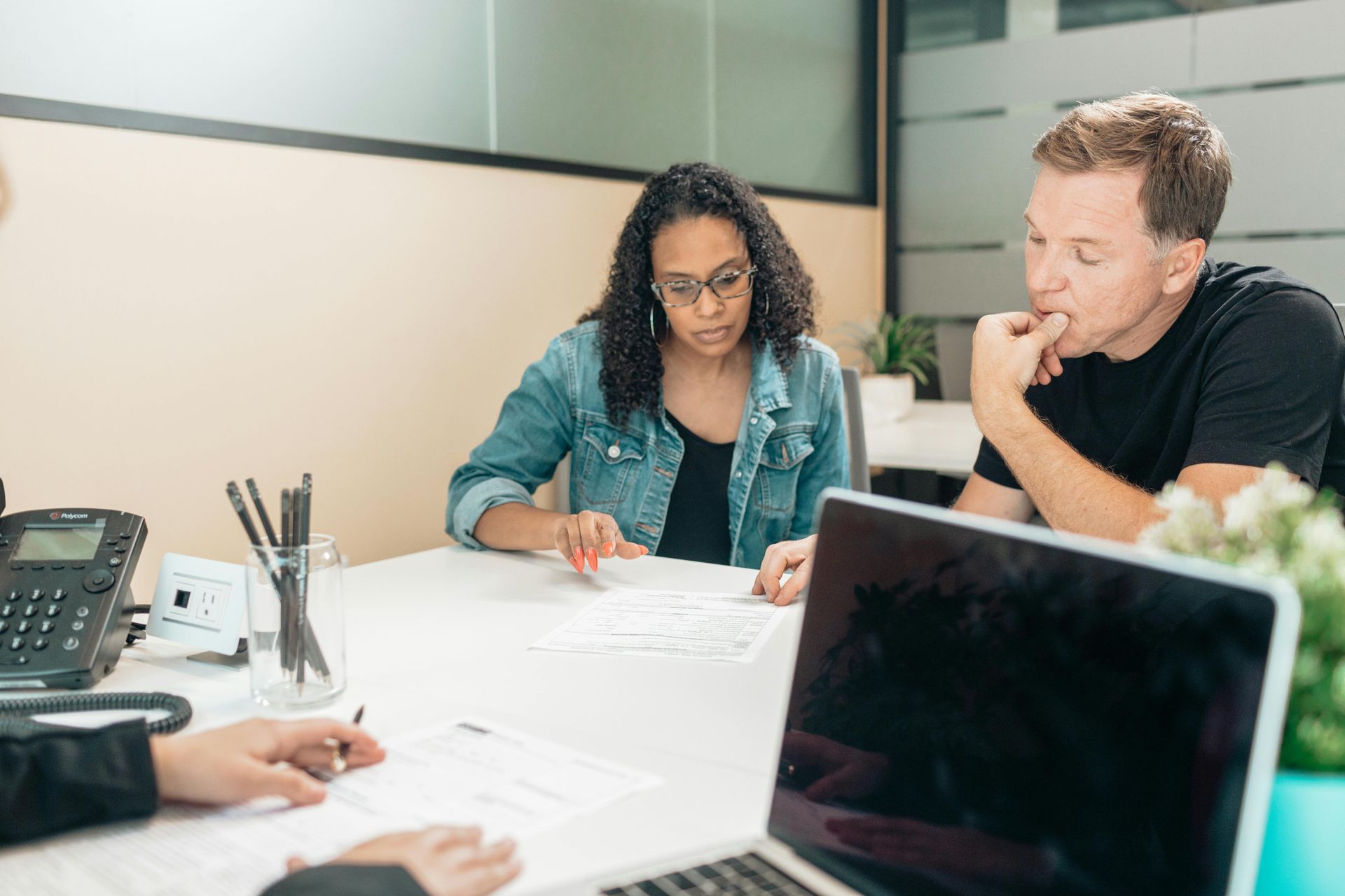 Three people sit at a desk in an office meeting, reviewing documents while talking.