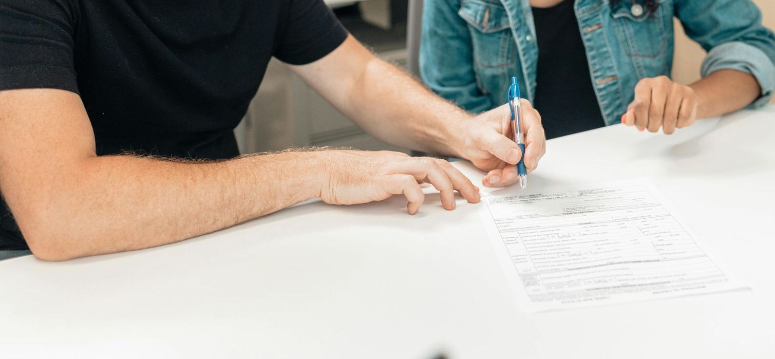 Two people sit at a white table, one signing a document with a blue pen while the other watches.