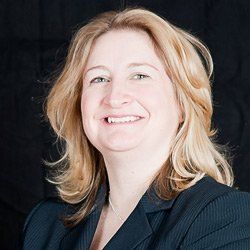 A person with shoulder-length blonde hair, wearing a dark blazer, smiling against a black background.