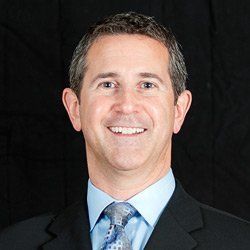 A smiling professional wearing a dark suit, light blue dress shirt, and patterned tie against a black background.