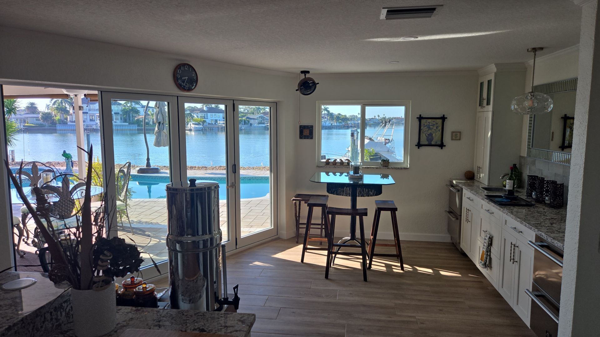 Bright kitchen with a view of a pool and waterway. White cabinets, bar with stools, and sliding glass doors.