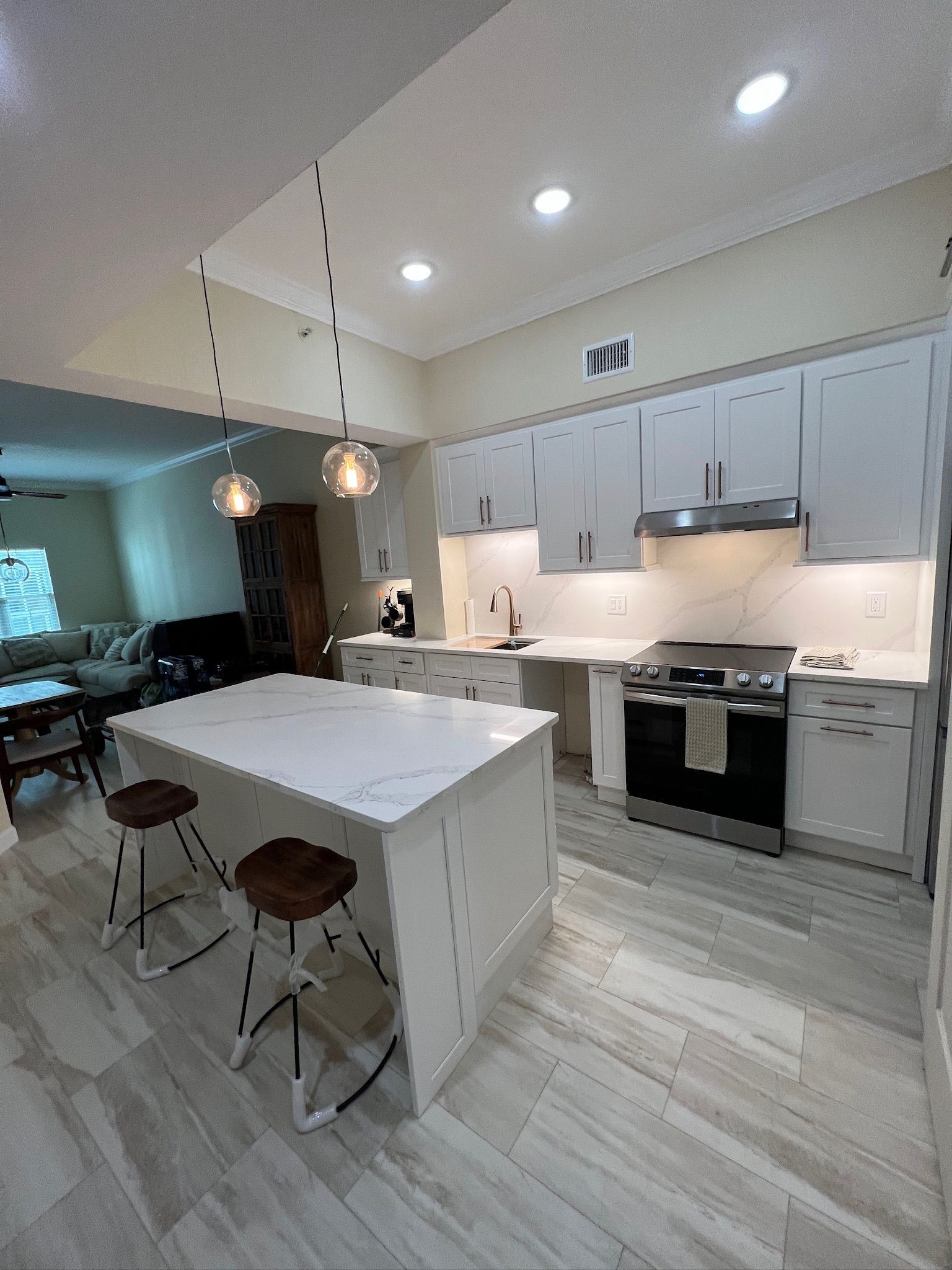 Bright modern kitchen with white cabinets, island, pendant light, and two stools, open to a living room.