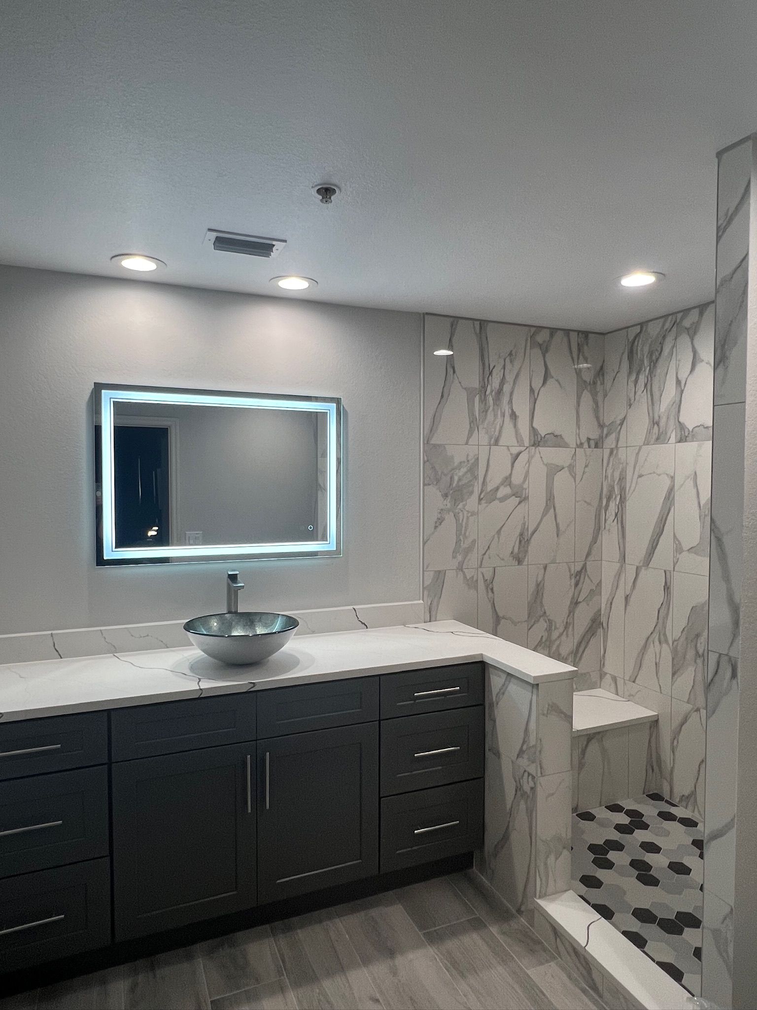 Modern bathroom vanity with lit mirror, dark cabinets, white countertop, and marble-patterned shower tiles
