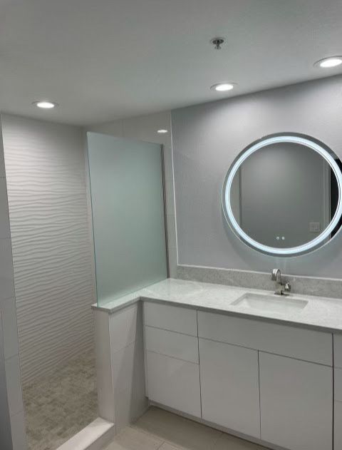 Modern bathroom vanity with a round lit mirror, white cabinets, and a frosted glass shower enclosure