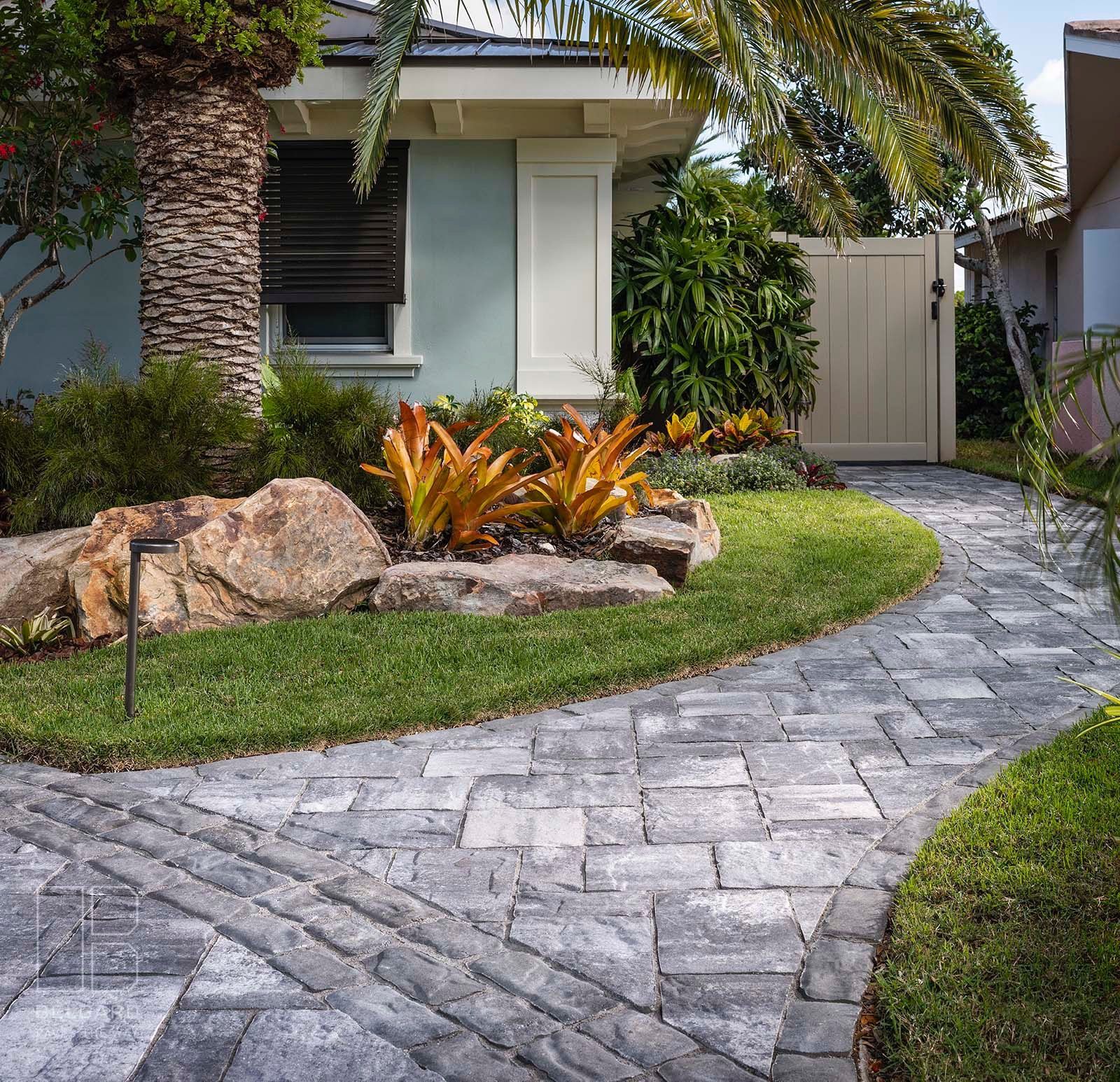 Curved stone walkway leading to a house, bordered by green lawn, rocks, and tropical plants