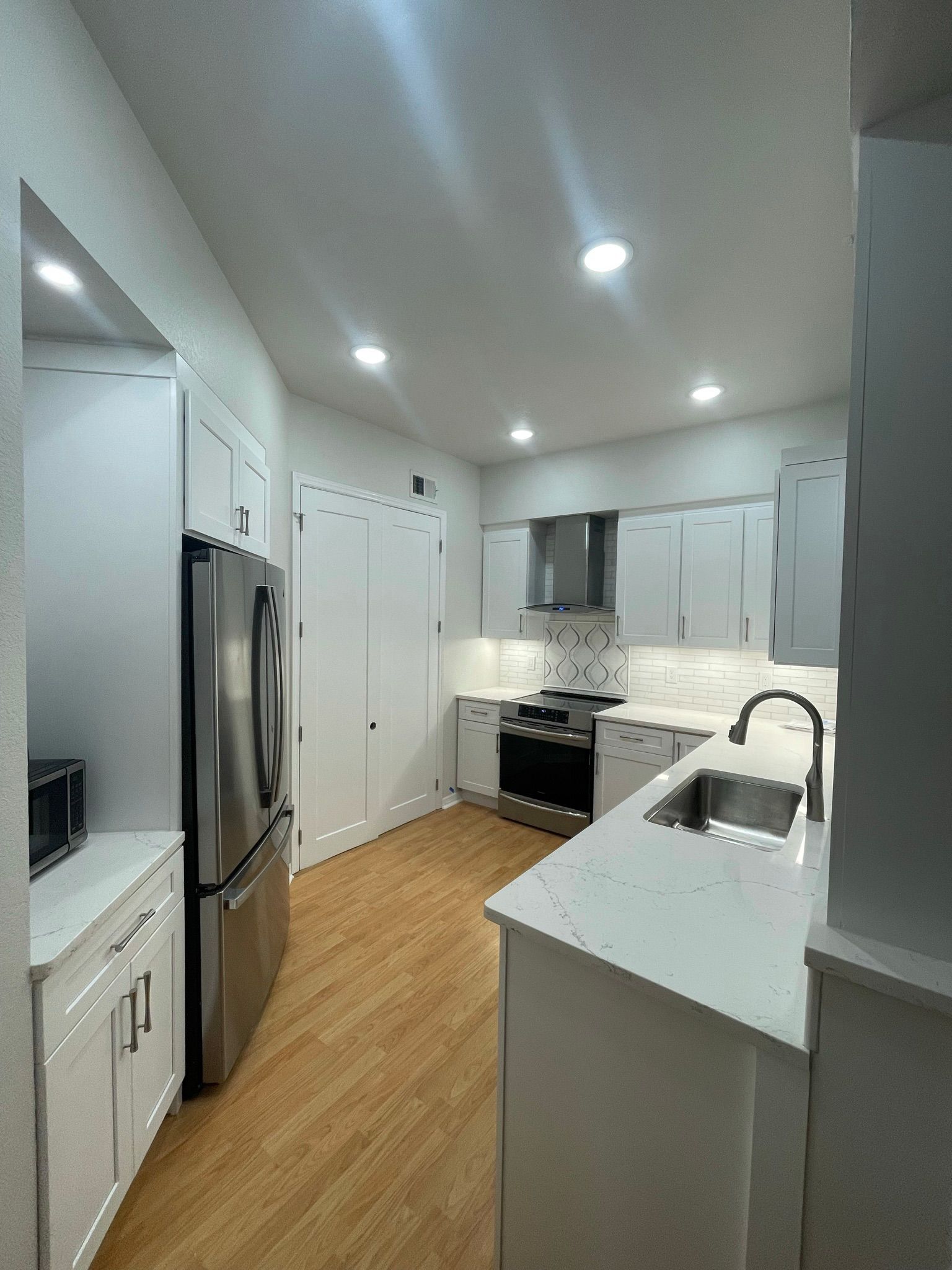 Modern white kitchen with island, stainless fridge, wood floor, and recessed ceiling lights