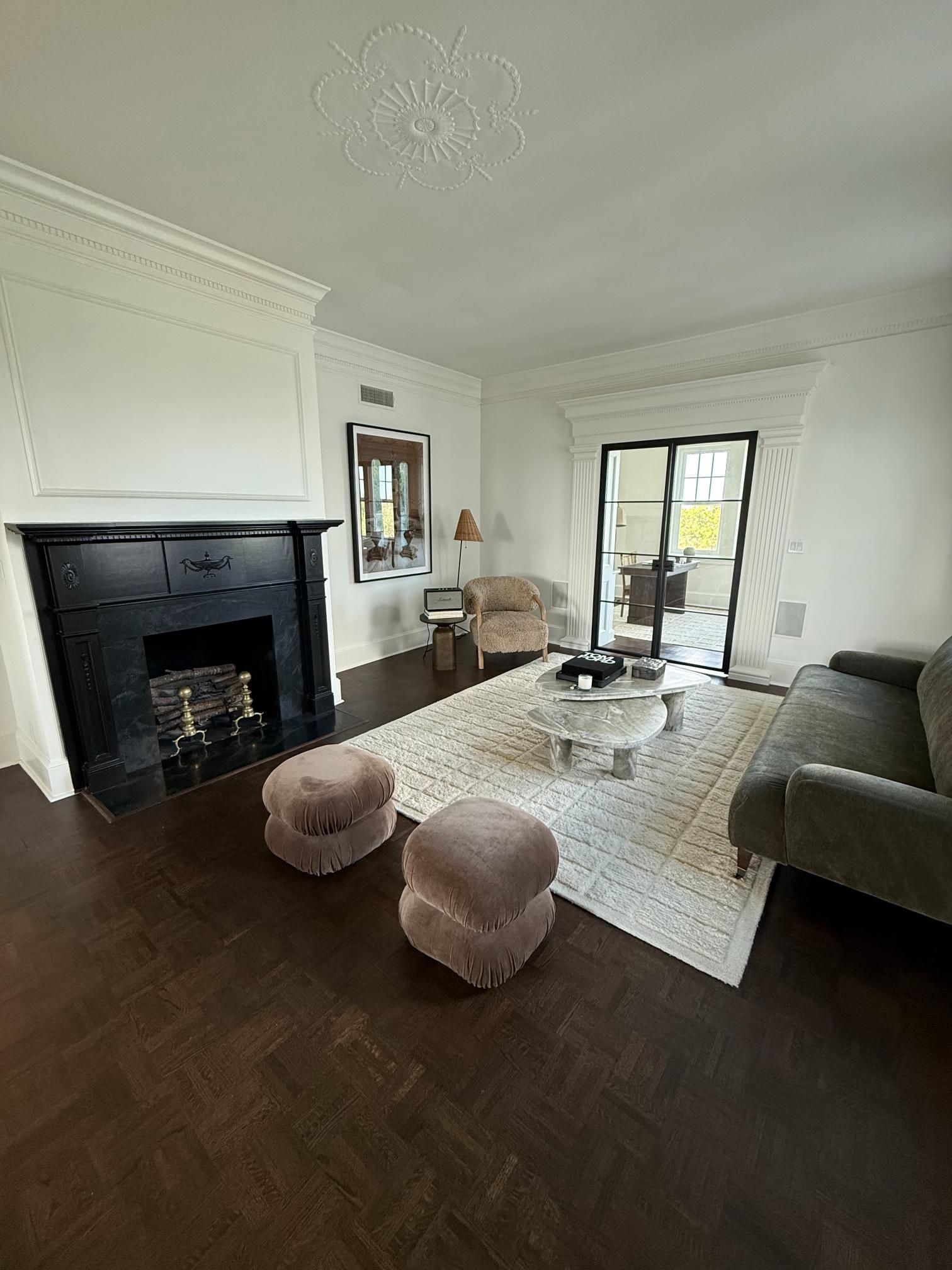 Bright living room with black fireplace, white walls, rug, poufs, and dark sofa by glass doors