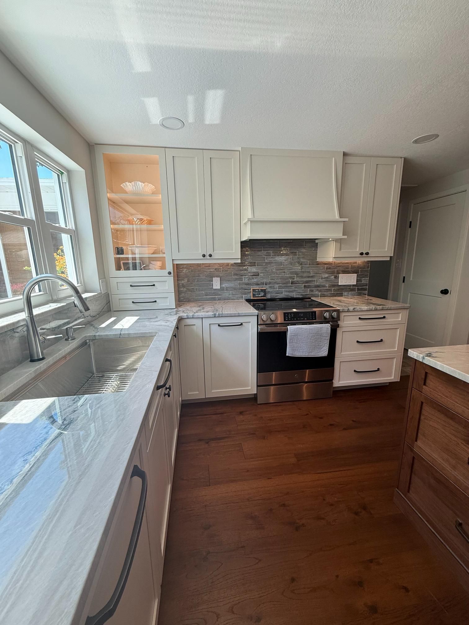 Bright modern kitchen with white cabinets, dark wood floor, large window, and stainless steel appliances