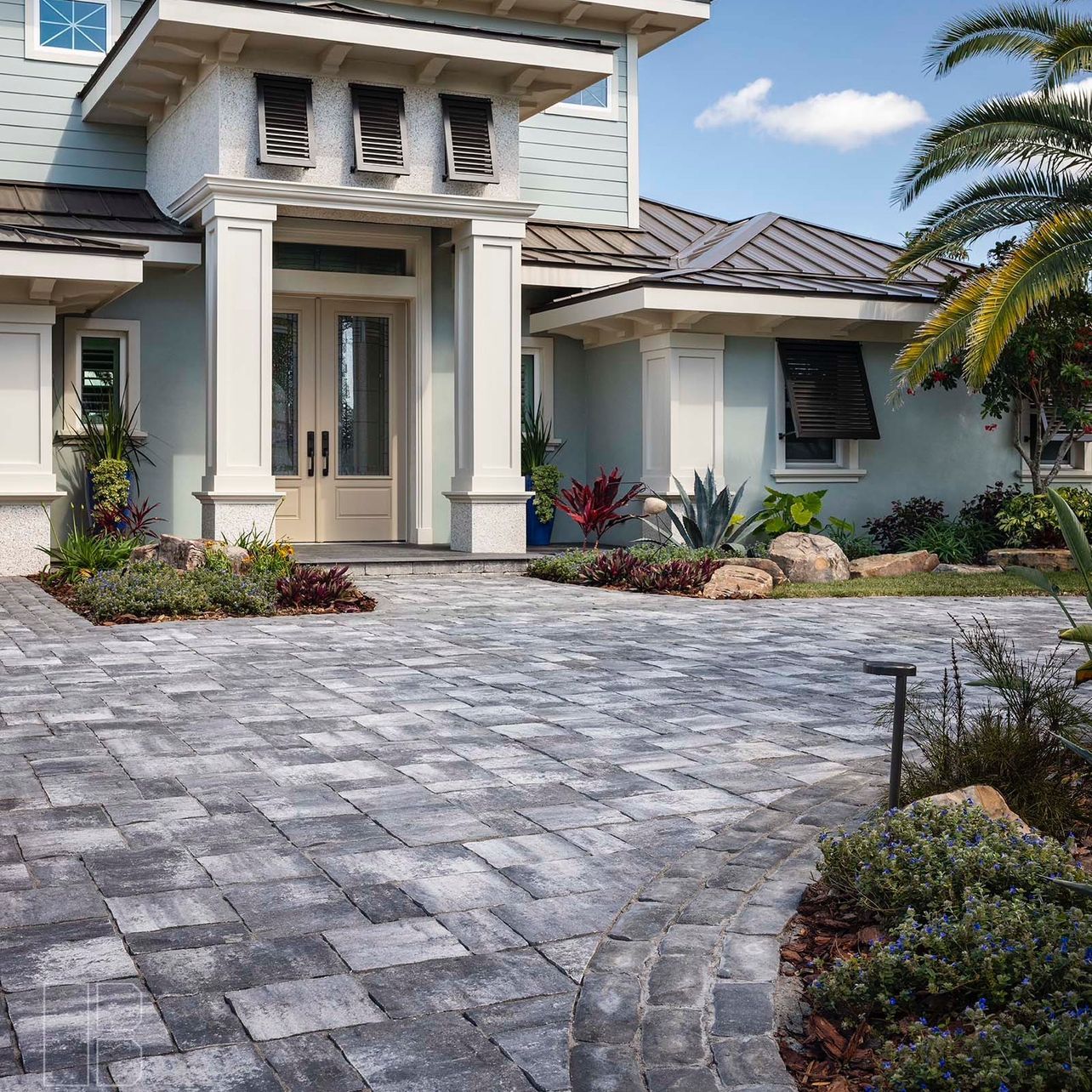 Modern house exterior with stone paver driveway, white columns, and tropical landscaping