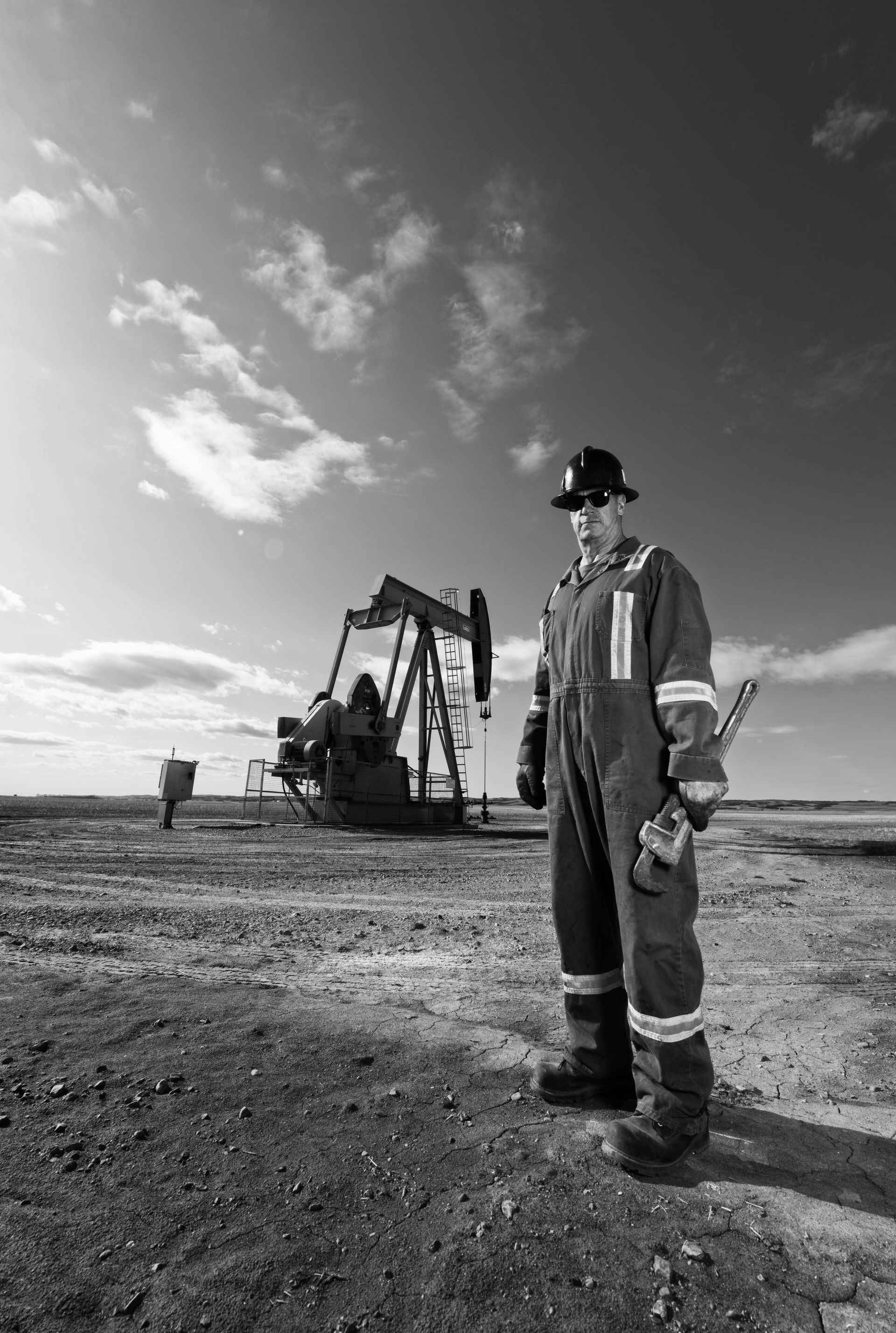 Oil worker in coveralls stands by a pump jack in a field under a sunny sky.