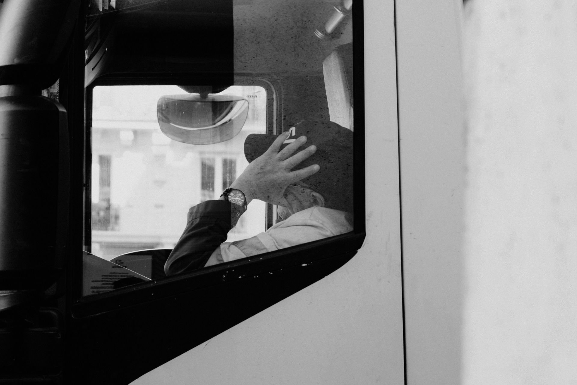 Person in driver's seat of vehicle, adjusting their hair. Seen from the side, through the window. Black and white.