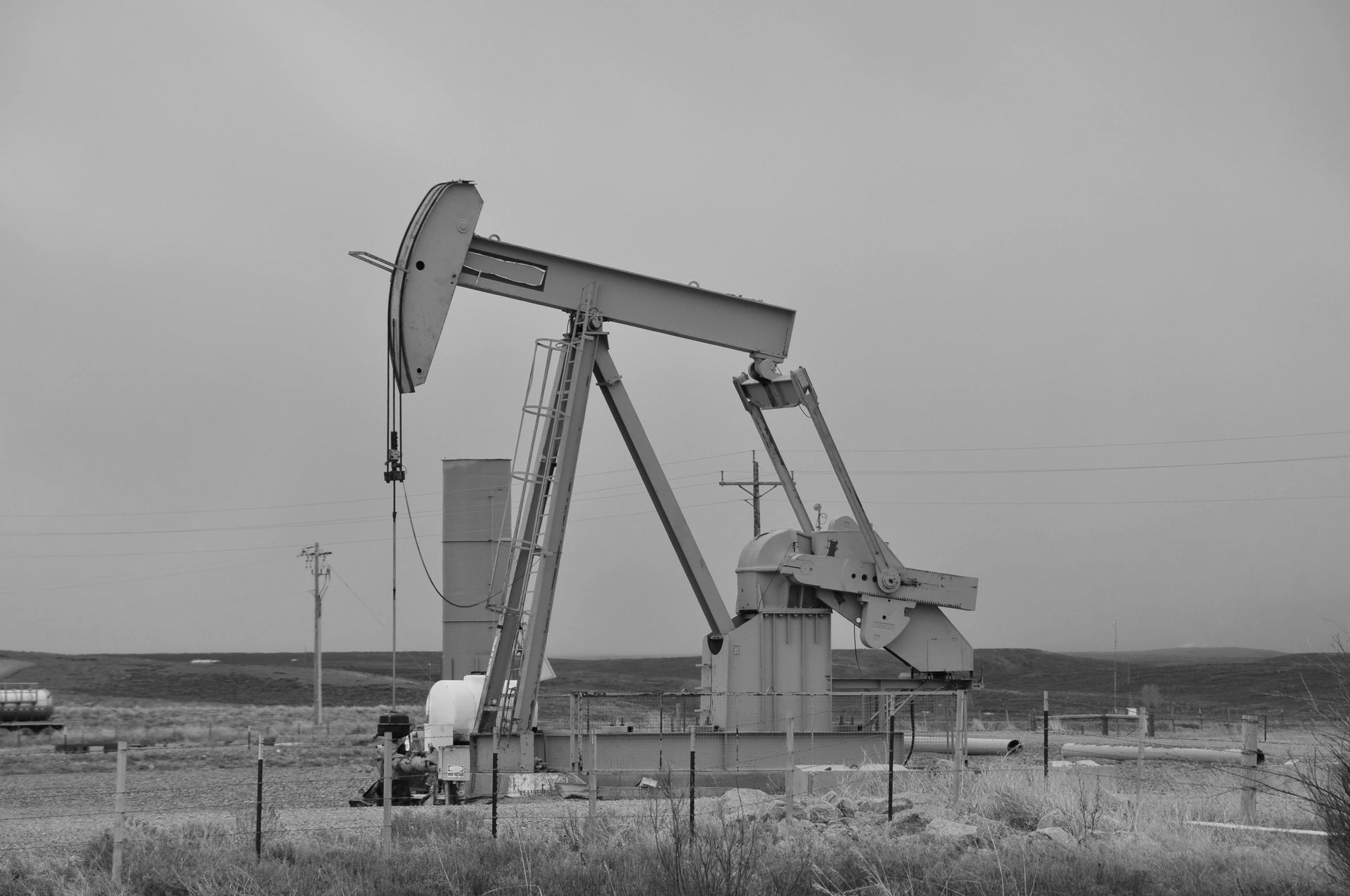 A grayscale image of an oil pump jack operating in a field, under a cloudy sky.