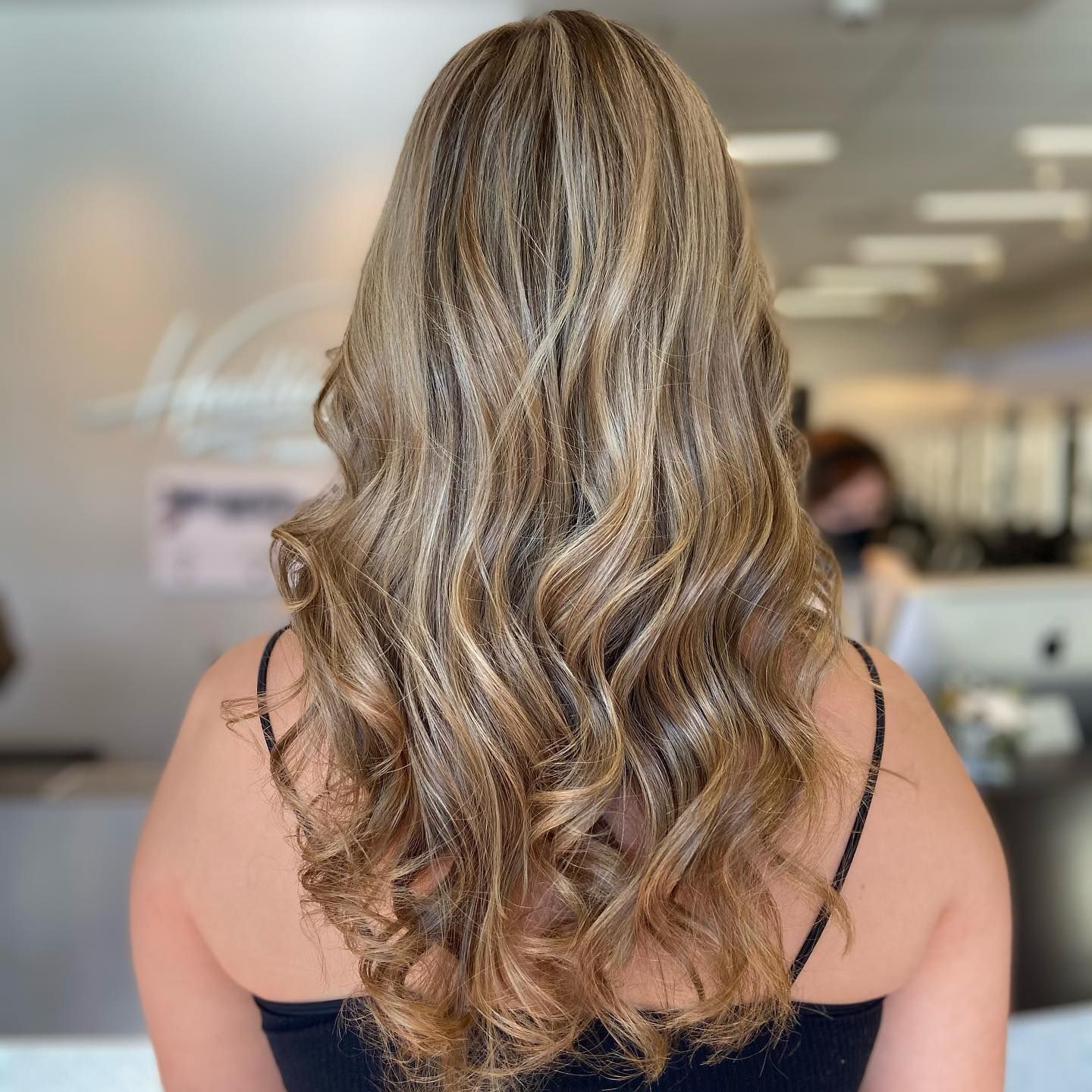 Woman with long, wavy, highlighted blonde hair, wearing a black top, in a salon.