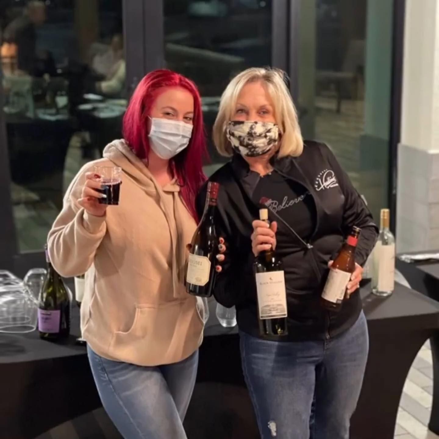 Two women in masks holding wine bottles and glasses at an event.