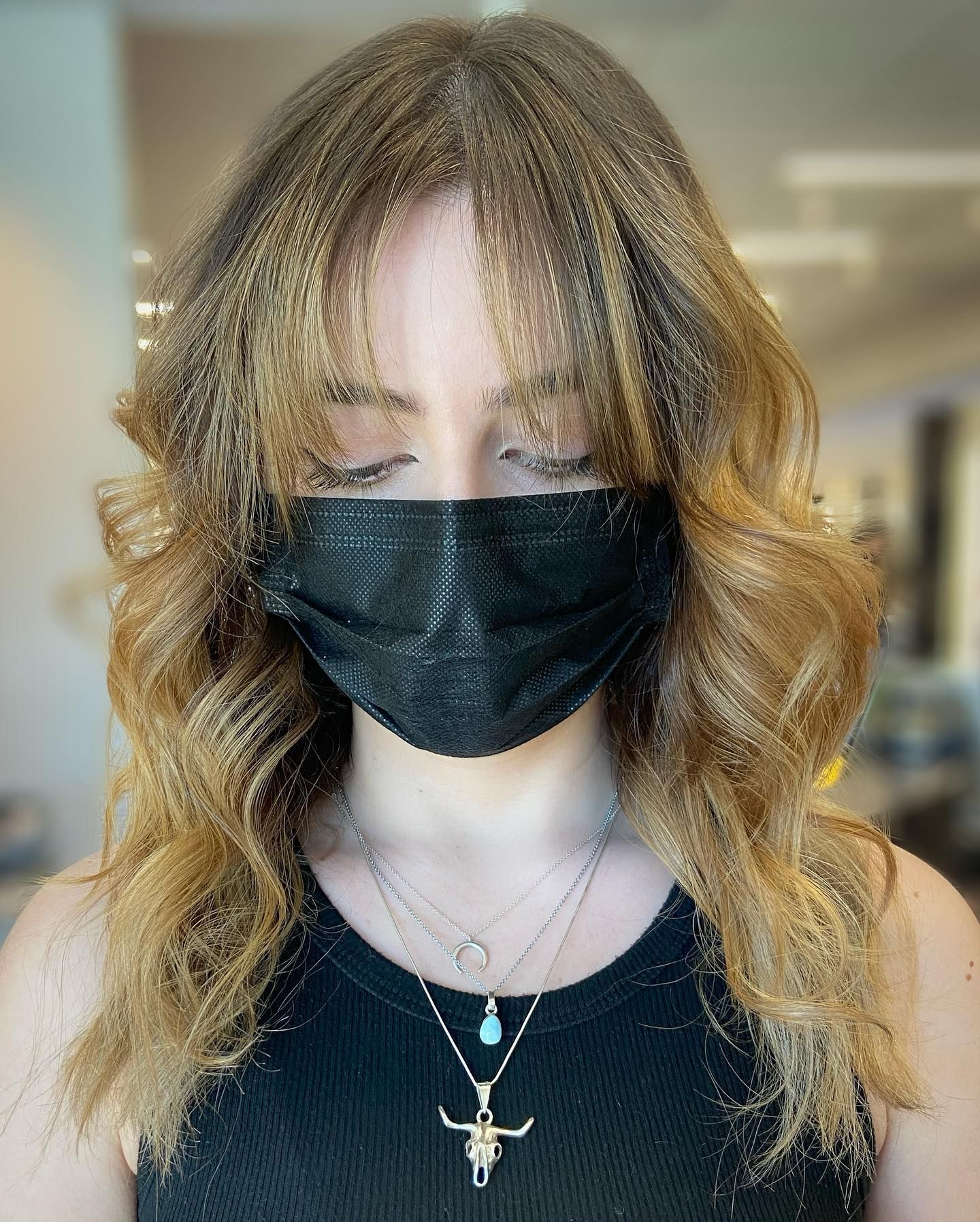Woman wearing a black face mask, with wavy blonde hair and layered bangs. She wears a black tank top and necklaces.