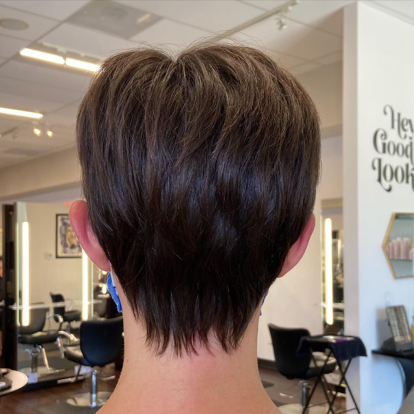 Back view of a person with short, dark brown layered hair, styled in a pixie cut, in a salon.