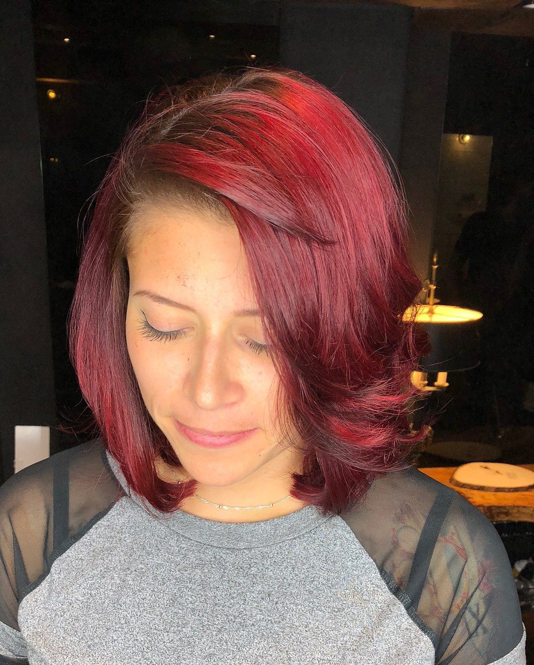 Woman with reddish-purple hair, looking down; wearing a gray shirt with black mesh; indoor setting.
