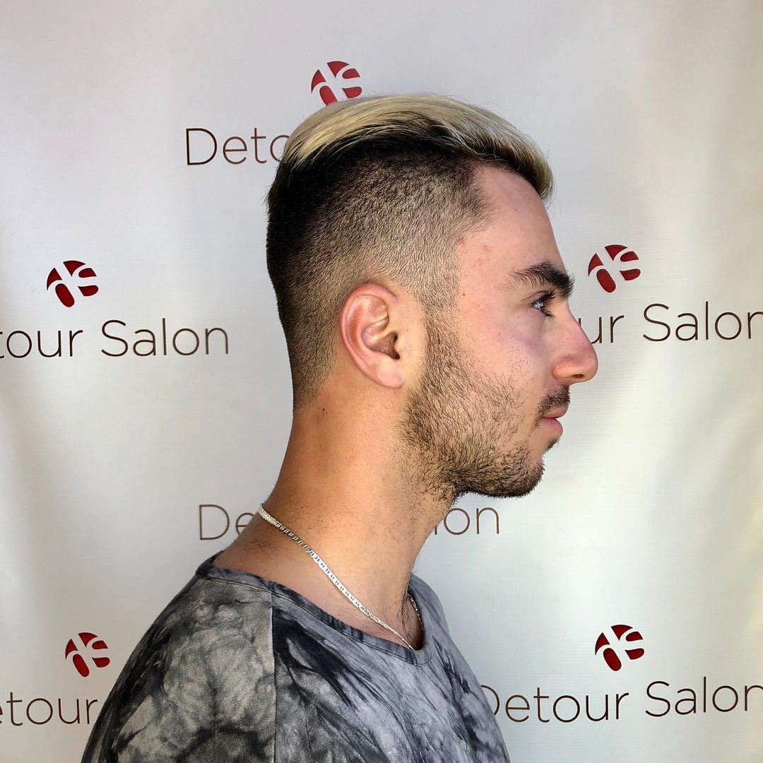 Man with blonde-tipped hair, short sides, and stubble. He is wearing a gray t-shirt, standing in front of a white wall.