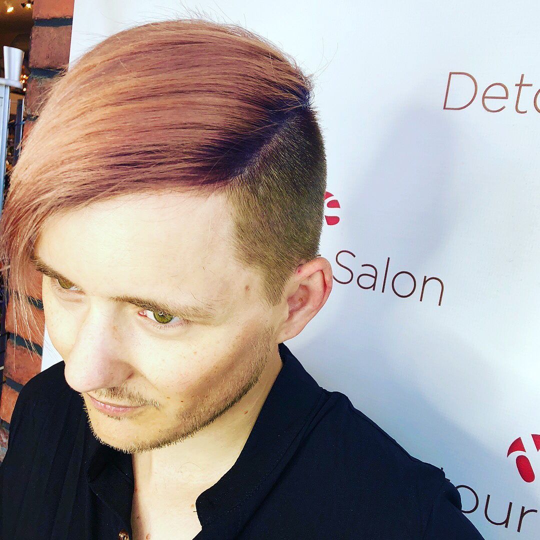 Man with pink hair and shaved sides, posing in front of a salon sign.