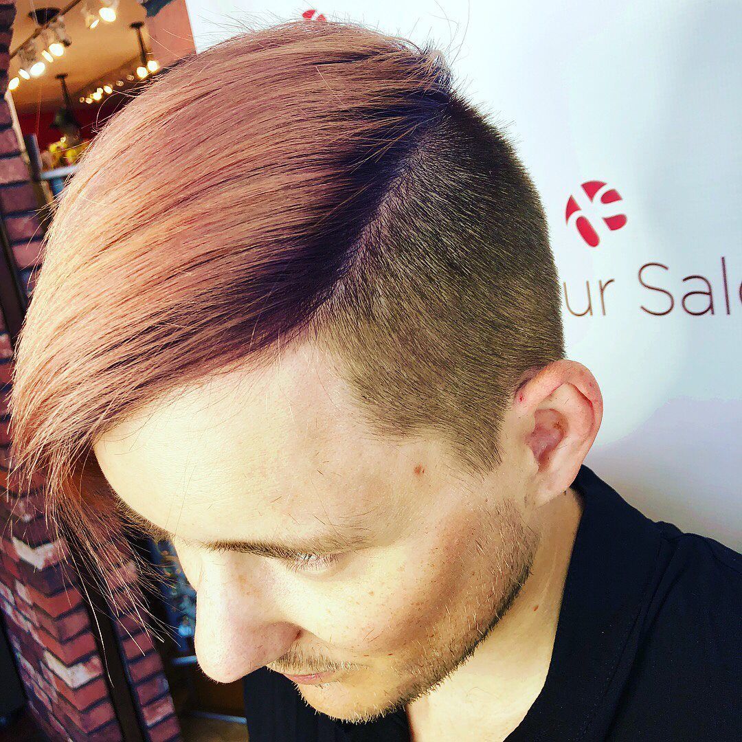 Man with angled side-swept pink hair and shaved sides, looking down. Brick wall and shop interior in background.