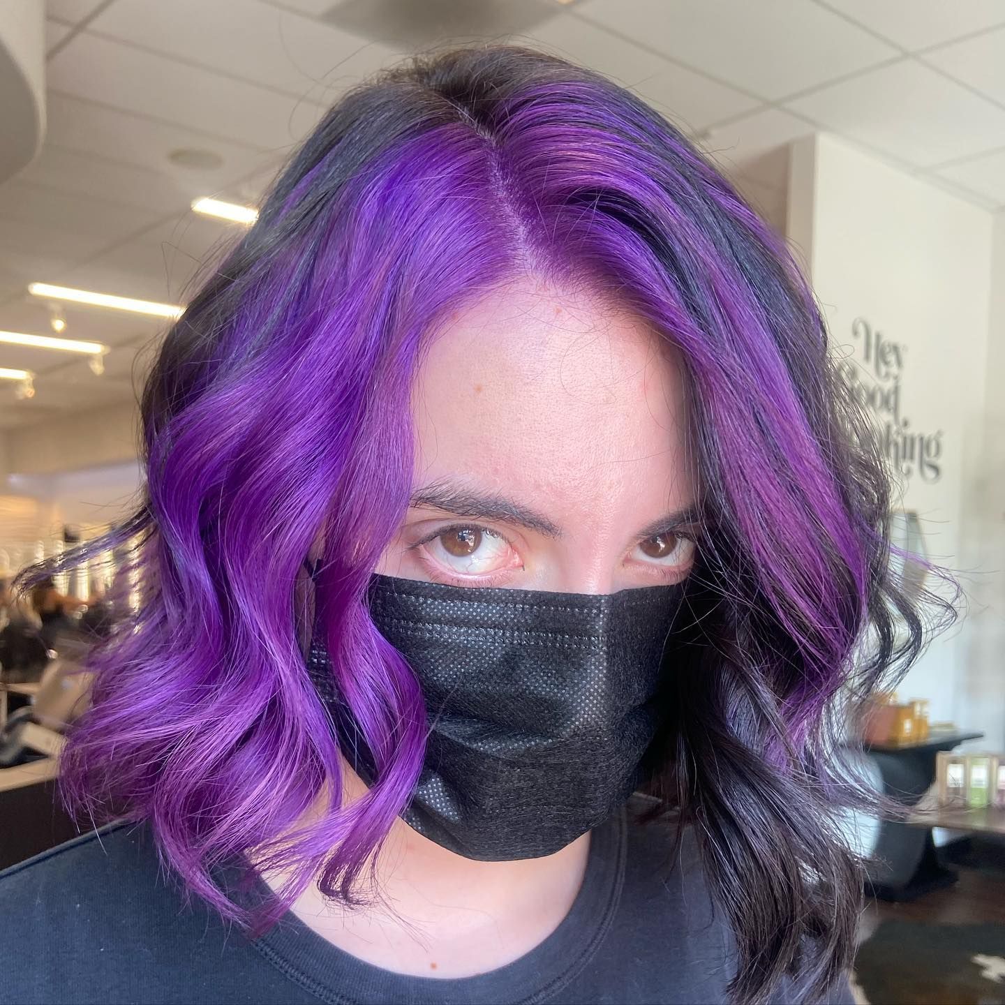Woman with purple and black wavy hair, wearing a black face mask, indoors.