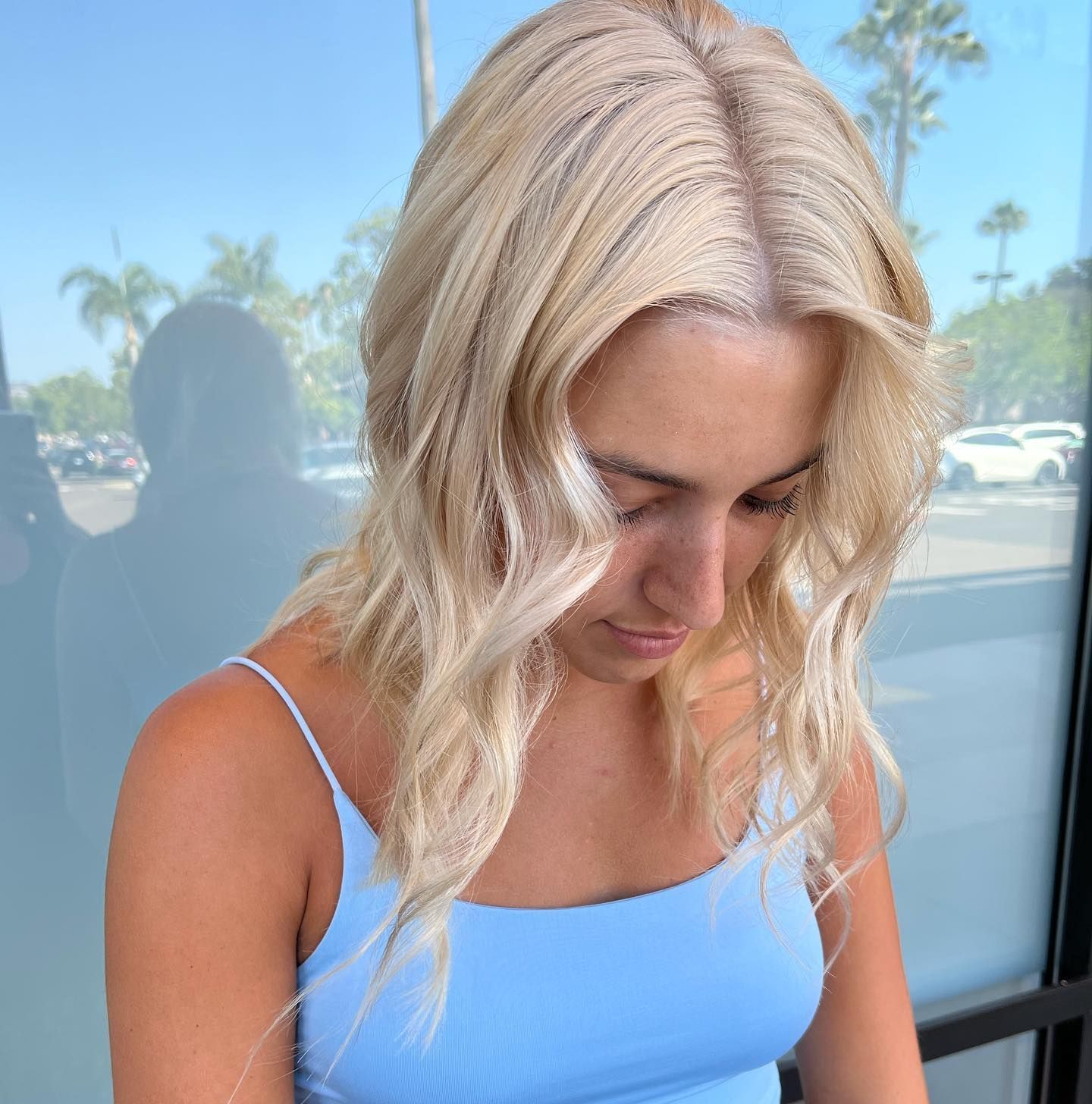 Woman with wavy blonde hair wearing a light blue tank top, looking down. Outdoors, sunny.