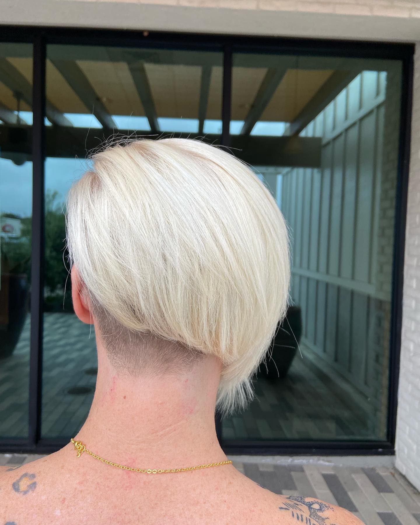 Person with short, bleached blonde hair, nape shaved, gold necklace, and a tattoo on the shoulder.