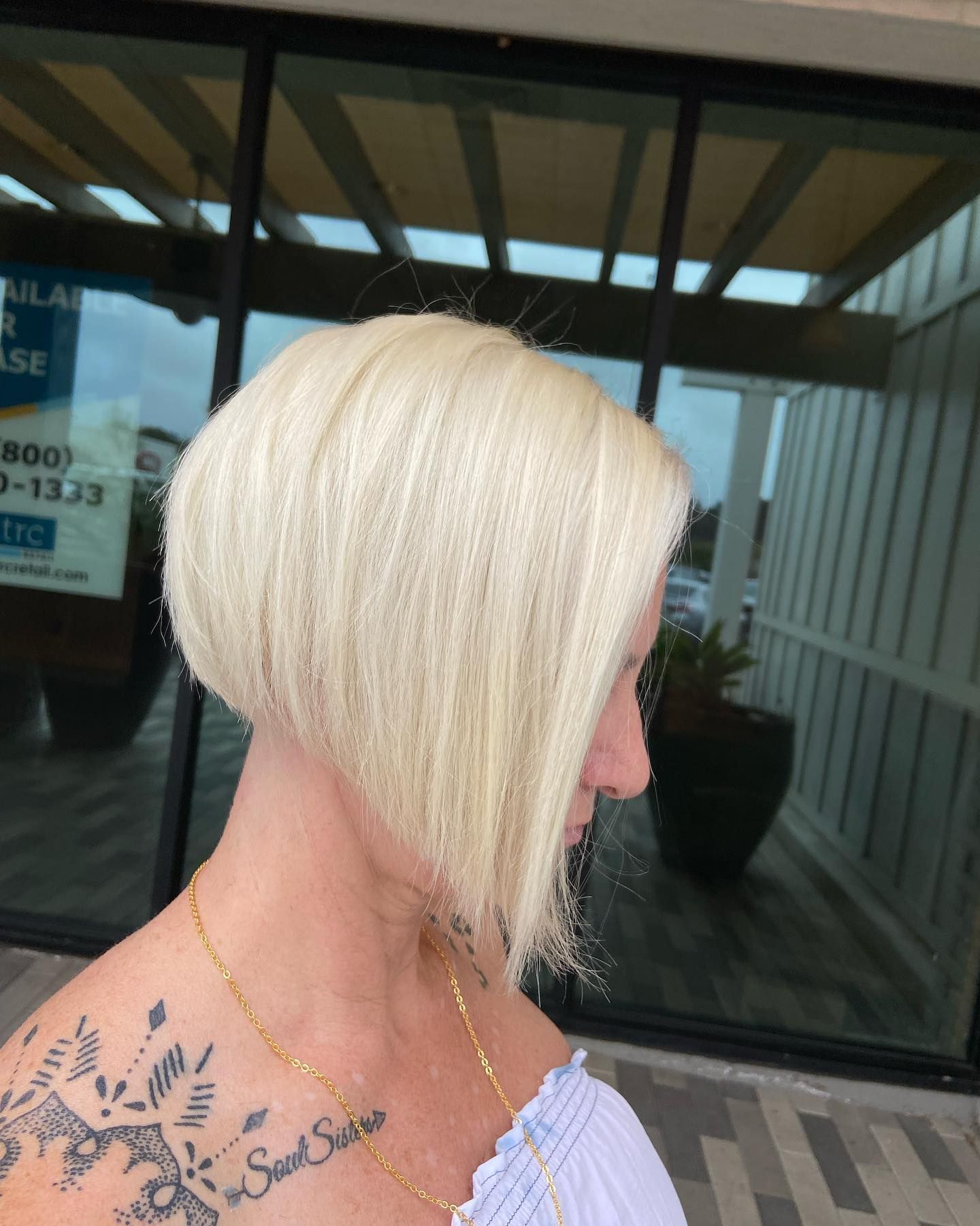 Woman with a blonde, asymmetrical bob haircut, wearing a white top, outside a building.