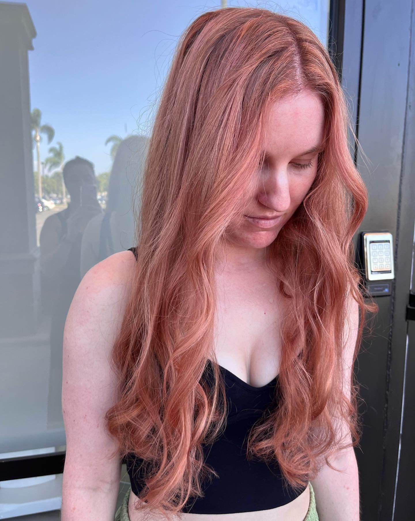 Woman with long, wavy auburn hair wearing a black top, looking down. Outdoors, sunny.