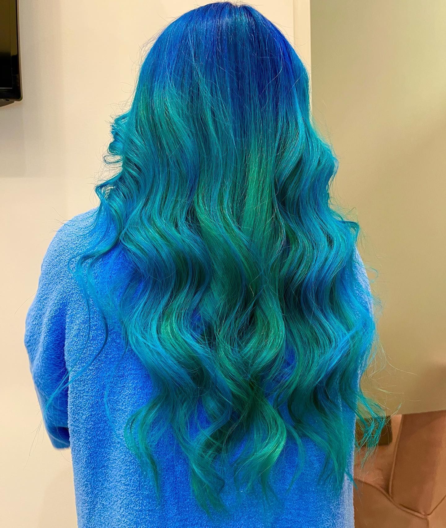 Person with long, wavy blue and teal hair, against a plain wall.