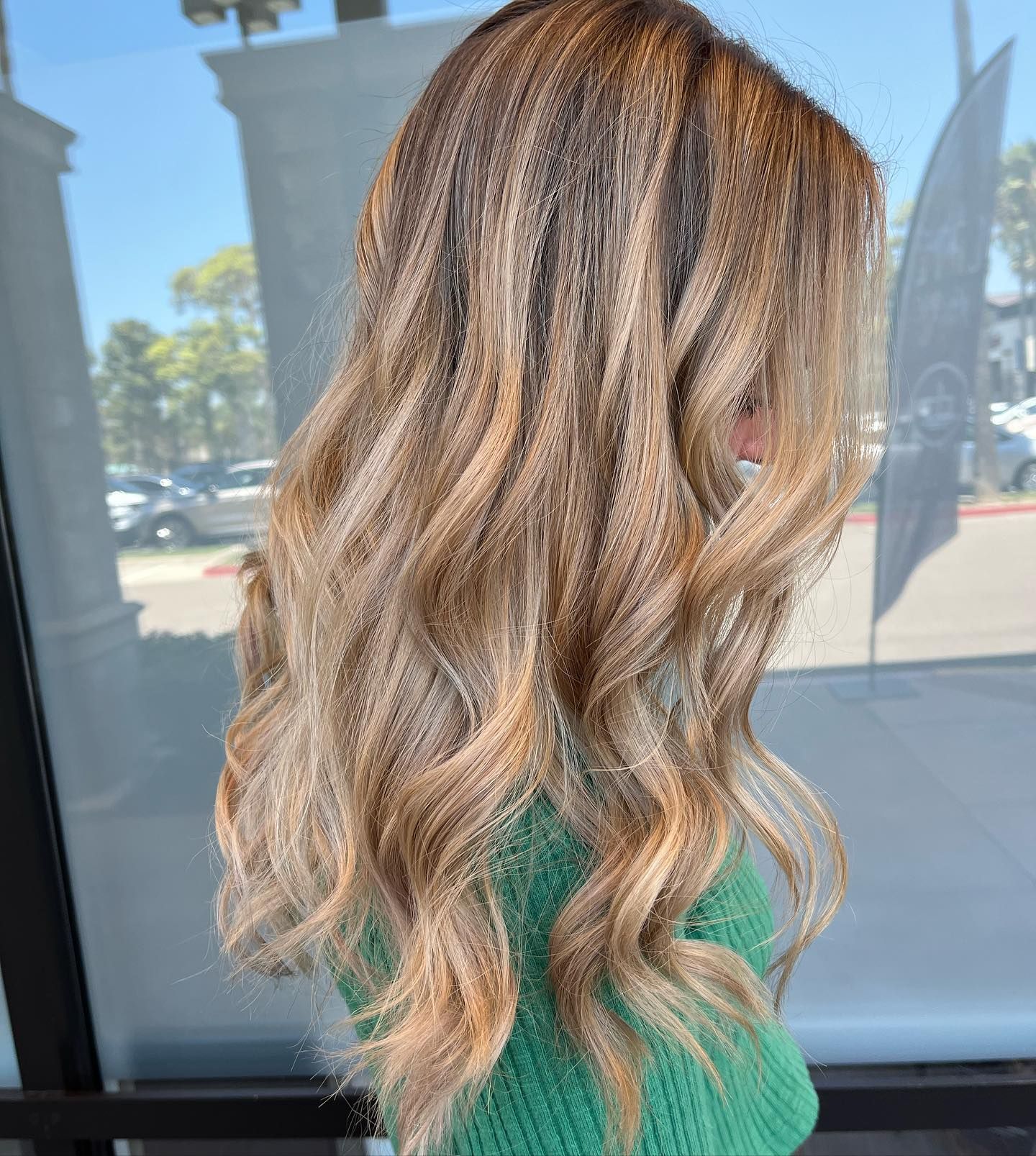 Woman with long, wavy blonde and brown highlighted hair; outside, wearing a green sweater.