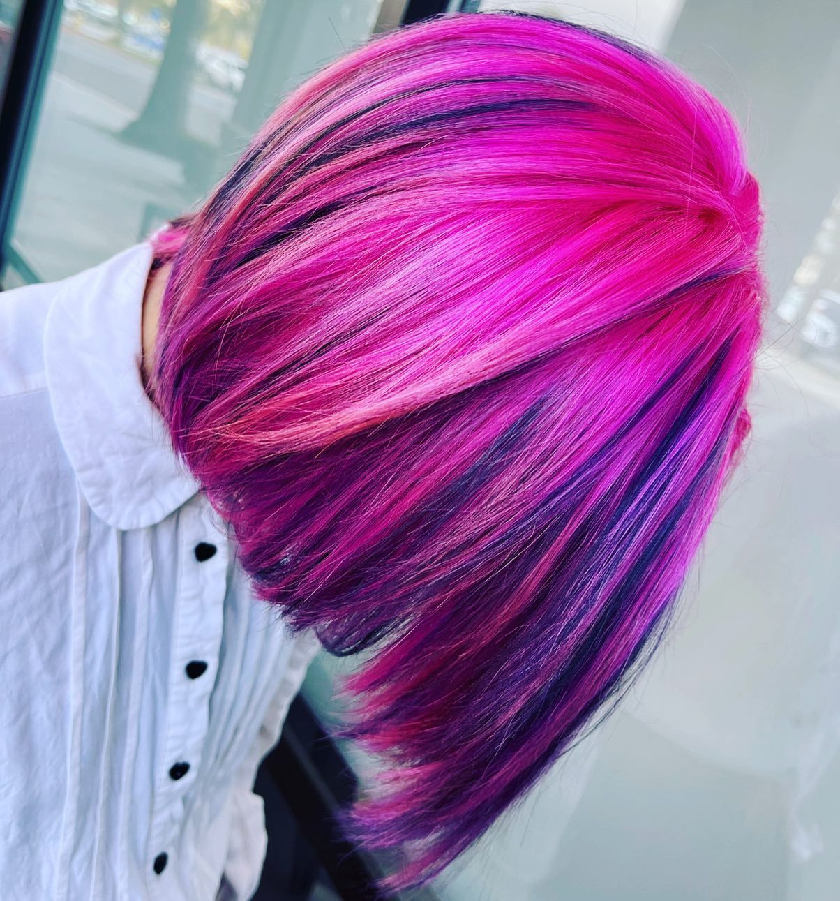 Pink and purple hair, bob style, in front of a white shirt, against a blurred background.