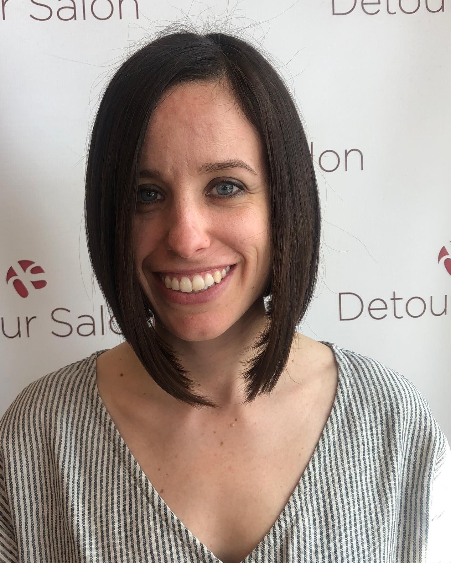 Woman with dark bob haircut smiling, wearing striped shirt, in a salon setting.