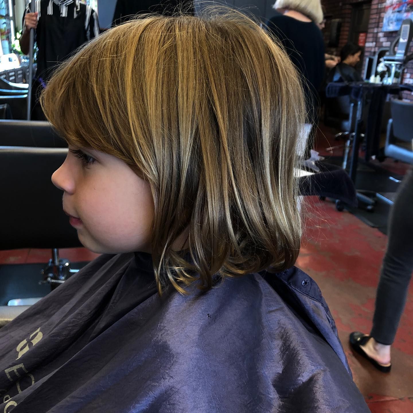 Child with layered, medium-length hair sits in a salon chair wearing a dark cape, side profile view.