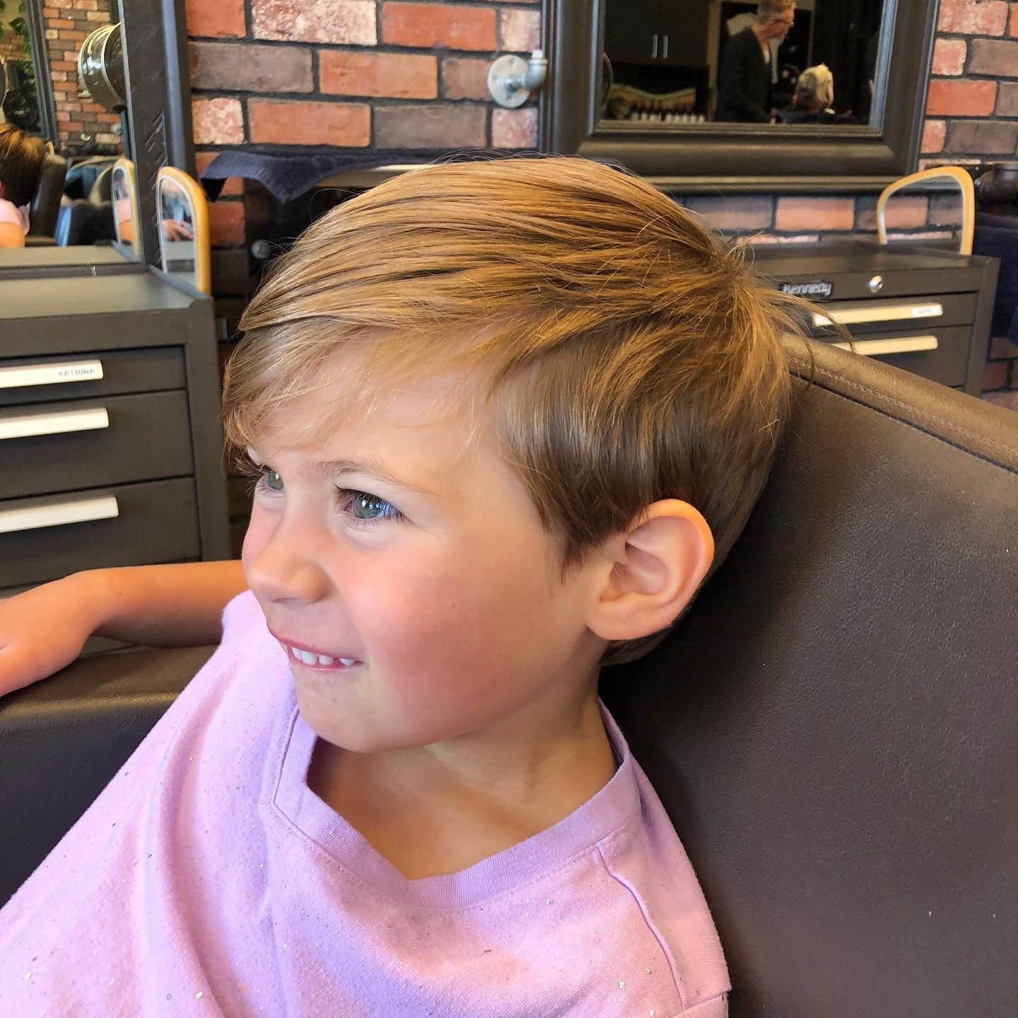Boy with blonde hair smiles, seated in a barber chair after a haircut.