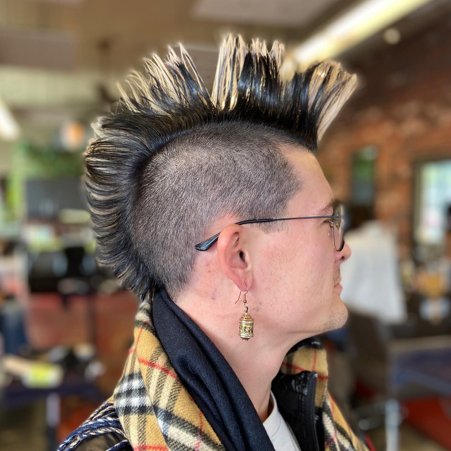 Man with mohawk hairstyle, glasses, and gold earring, wearing a patterned jacket in a barber shop.