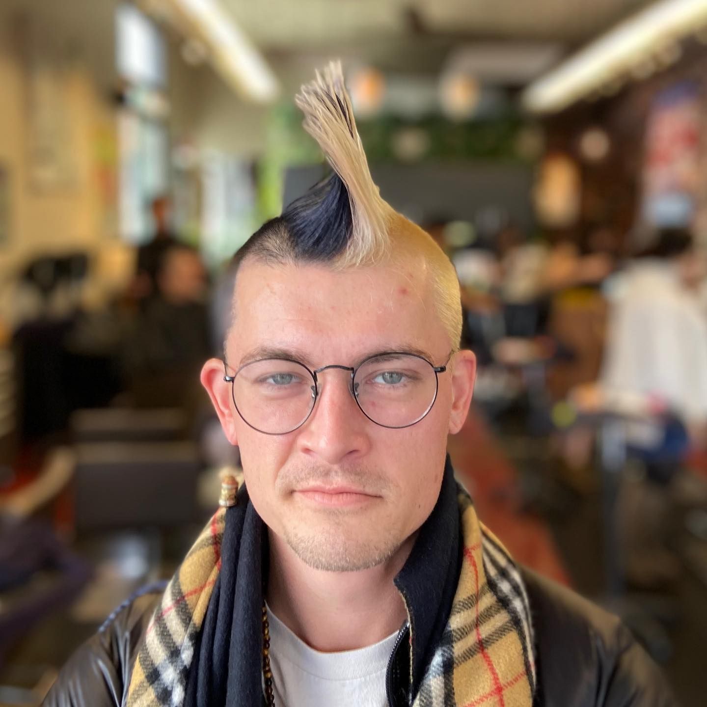 Person with a spiky, two-toned hairstyle and glasses, in a salon. Wearing a scarf and black jacket.