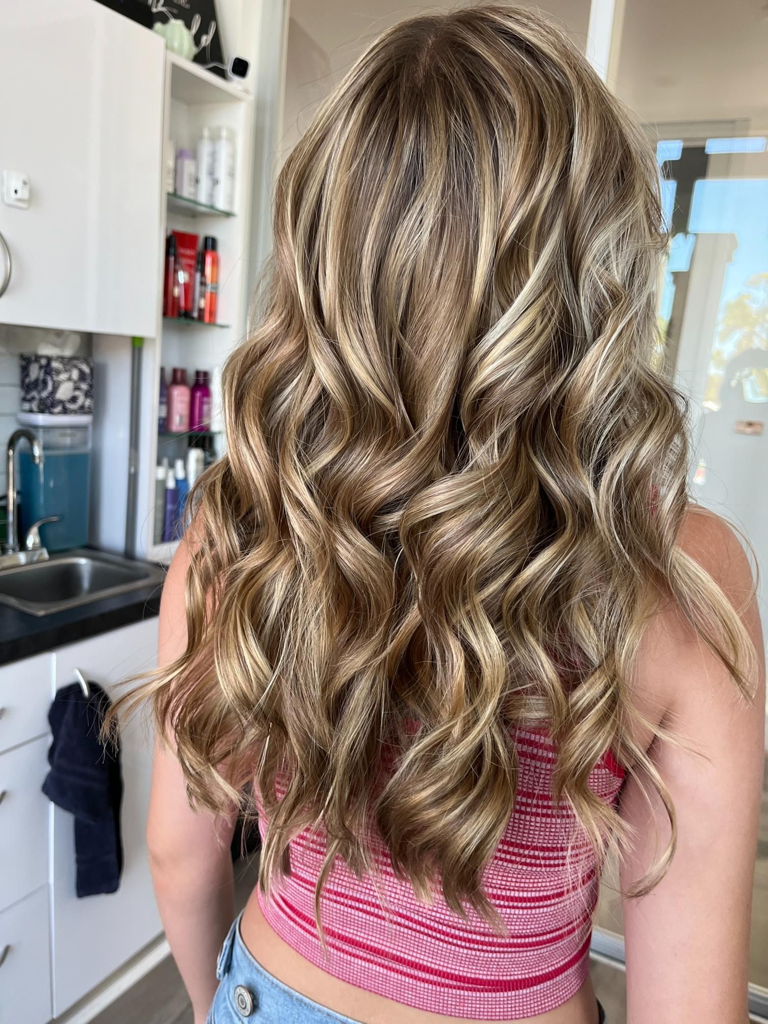 Woman with wavy, highlighted hair; wearing a red striped top. Hair salon setting.