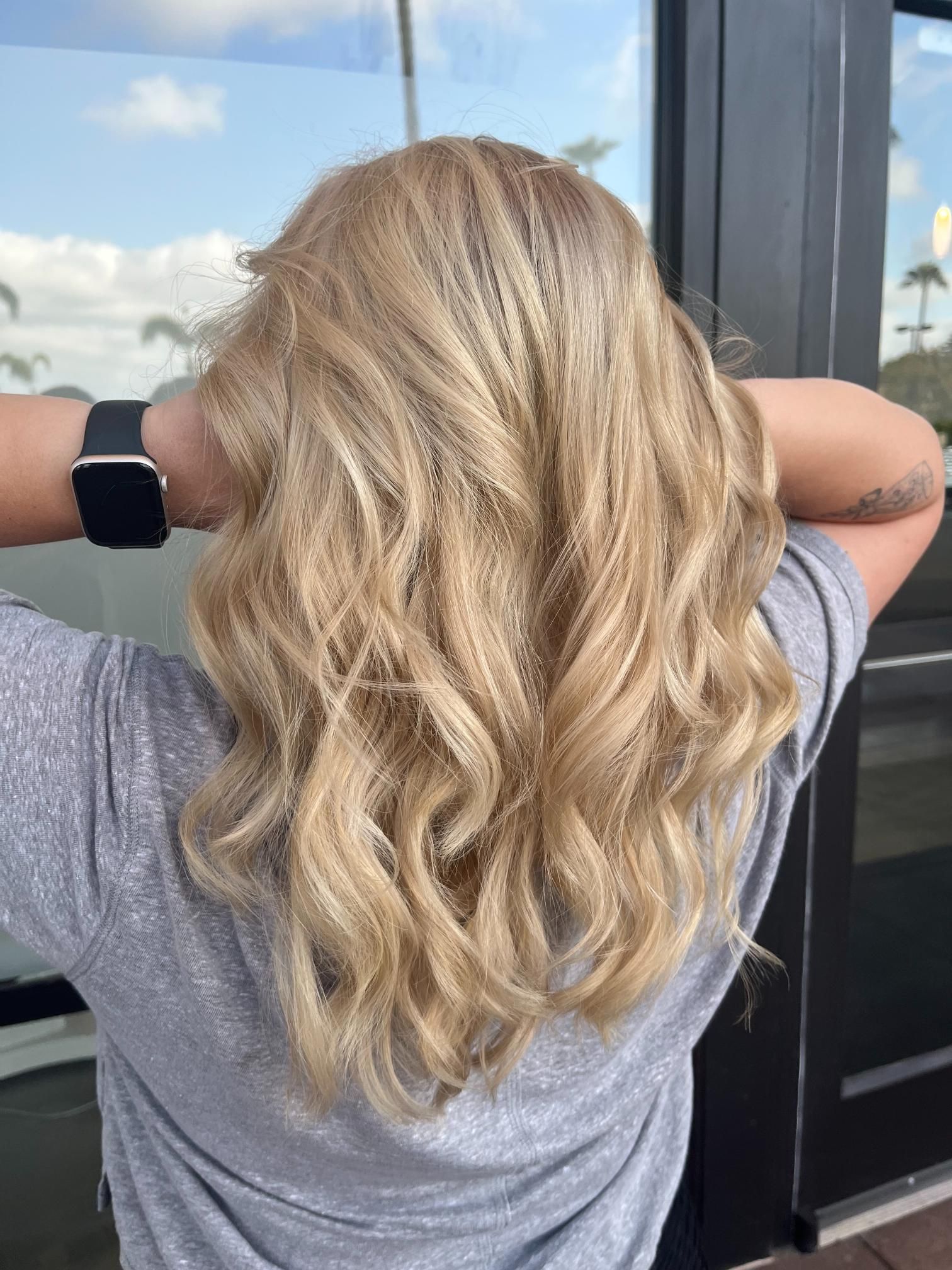 Woman with wavy, blonde hair, arms raised behind head. Wearing a gray shirt and smartwatch.