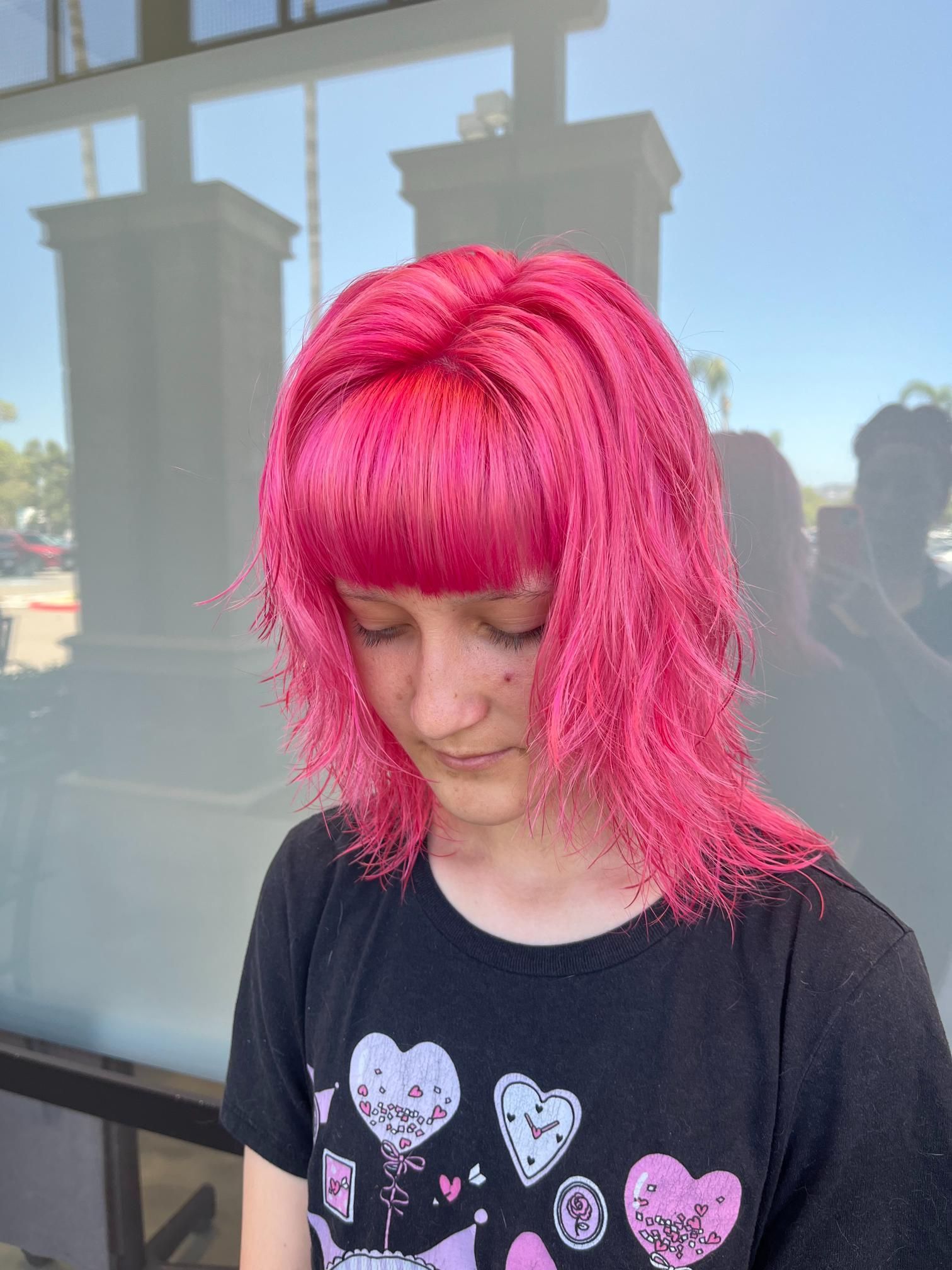 Person with pink hair and bangs wearing a black t-shirt, standing outdoors in front of a glass wall, looking down.