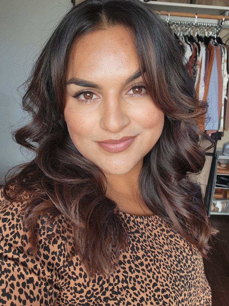 Woman with dark wavy hair and a leopard-print top, smiling at the camera. Neutral makeup. Clothes rack in background.