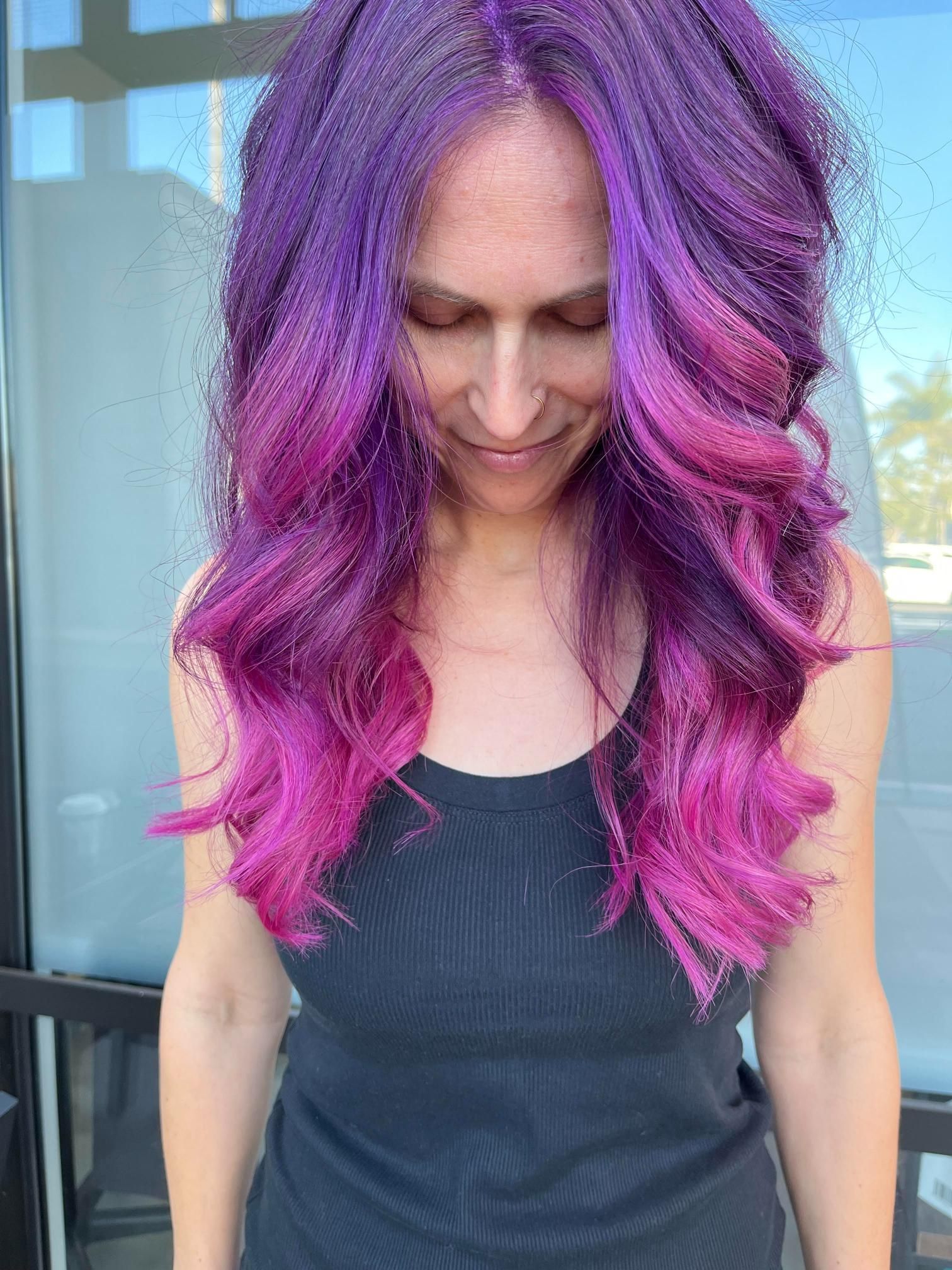 Woman with long, wavy purple and pink hair, looking down, wearing a black tank top.