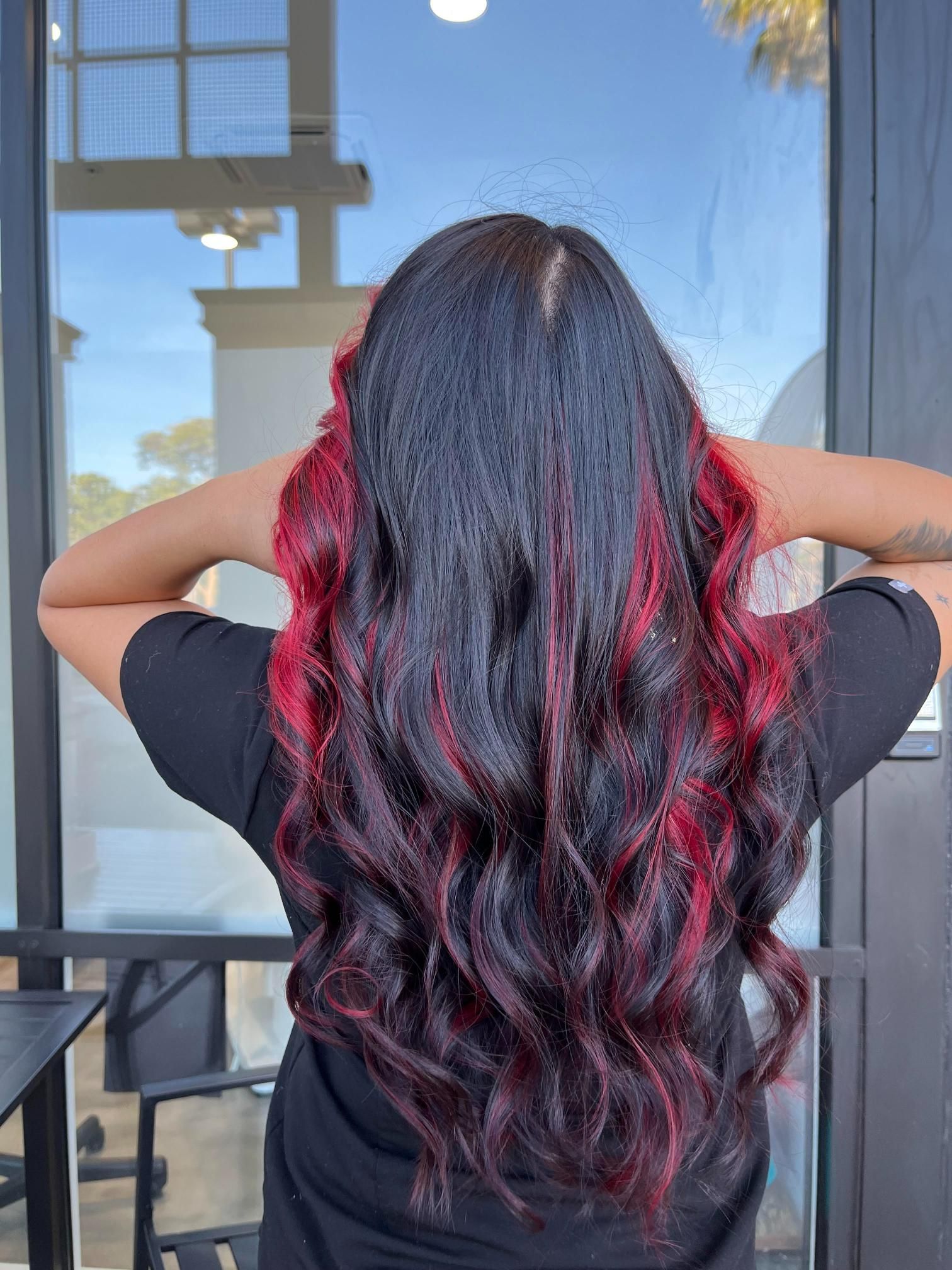 Person with long, wavy black hair with vibrant red highlights, standing in front of a glass door.