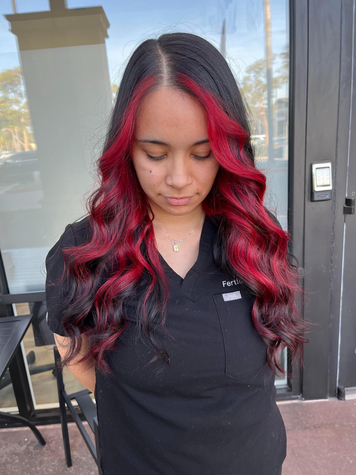 Woman with long black hair, red highlights, and wavy style, wearing a black shirt, outside.