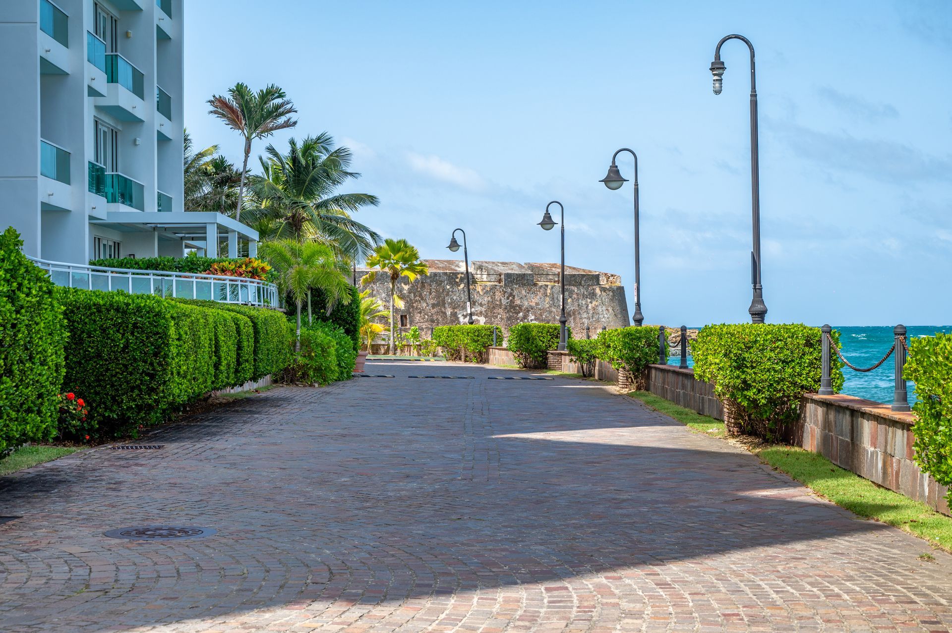 Seaside promenade with cobblestone path, hedges, palm trees, lamp posts, and ocean view beside a white building