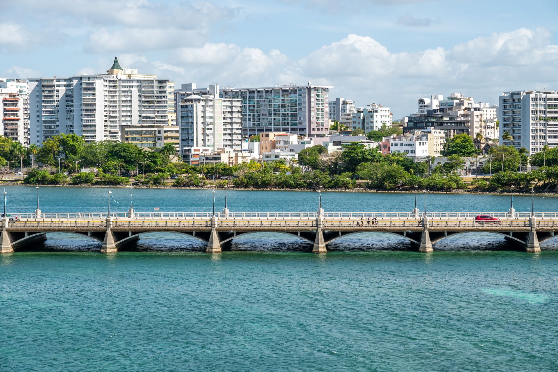 Waterfront bridge over turquoise water with a city skyline and white apartment buildings in the background