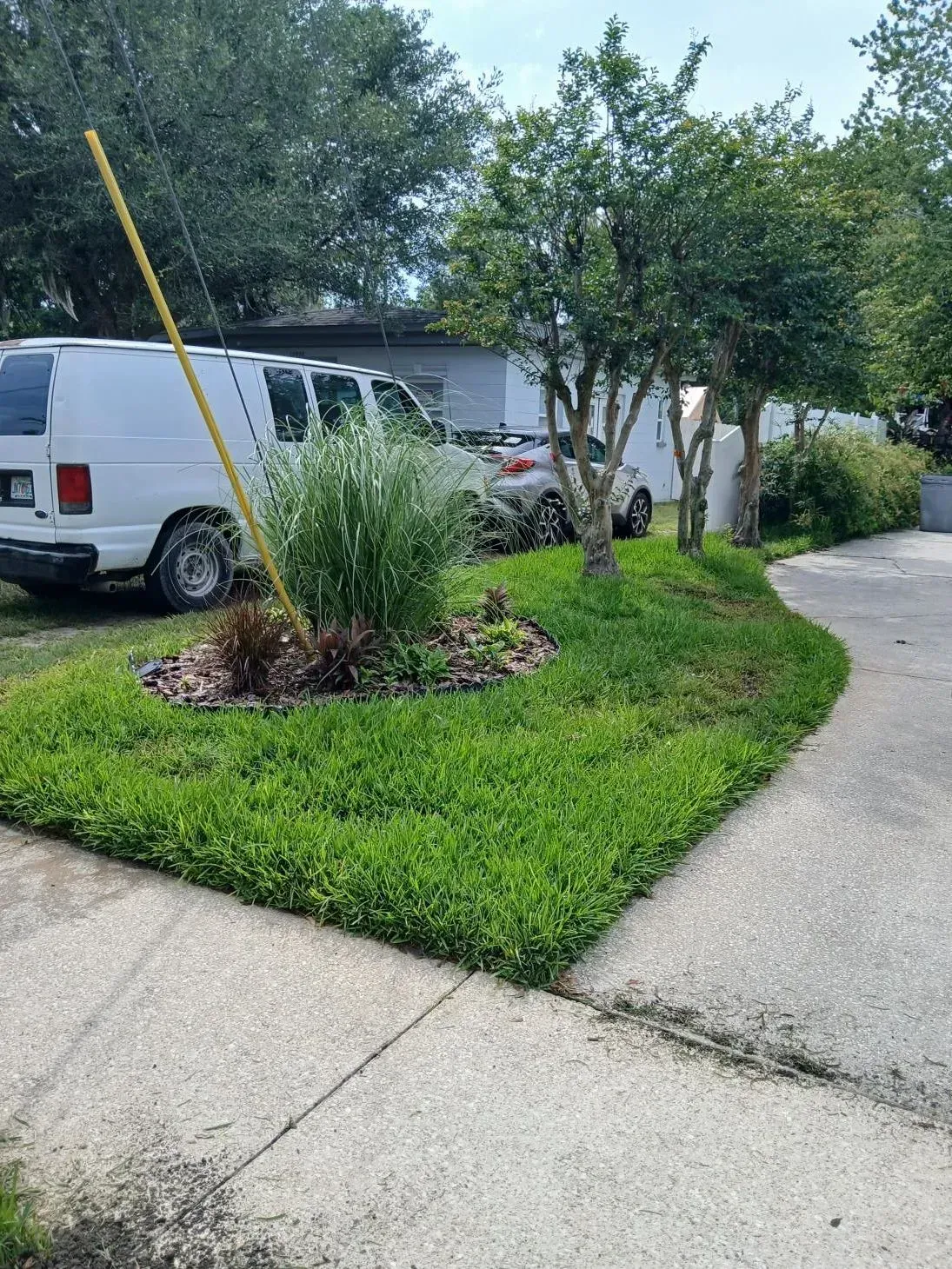 Suburban driveway beside a white van, trees, and a landscaped grassy corner with a yellow pole.